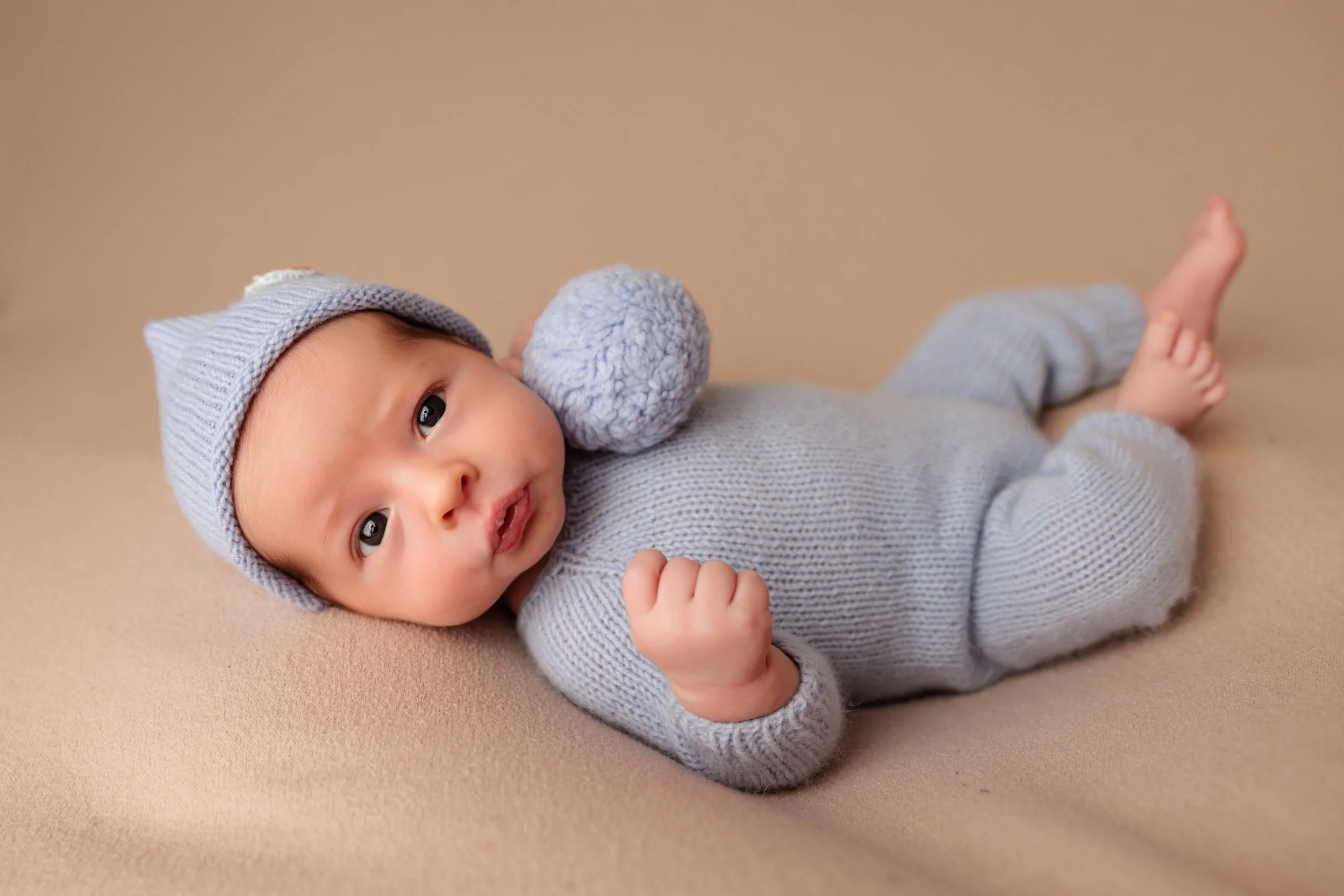 A baby dressed in a gray knitted outfit lying on a beige surface, wearing a matching gray hat, with a gray knitted pom-pom on their shoulder, looking at the camera with one fist raised.