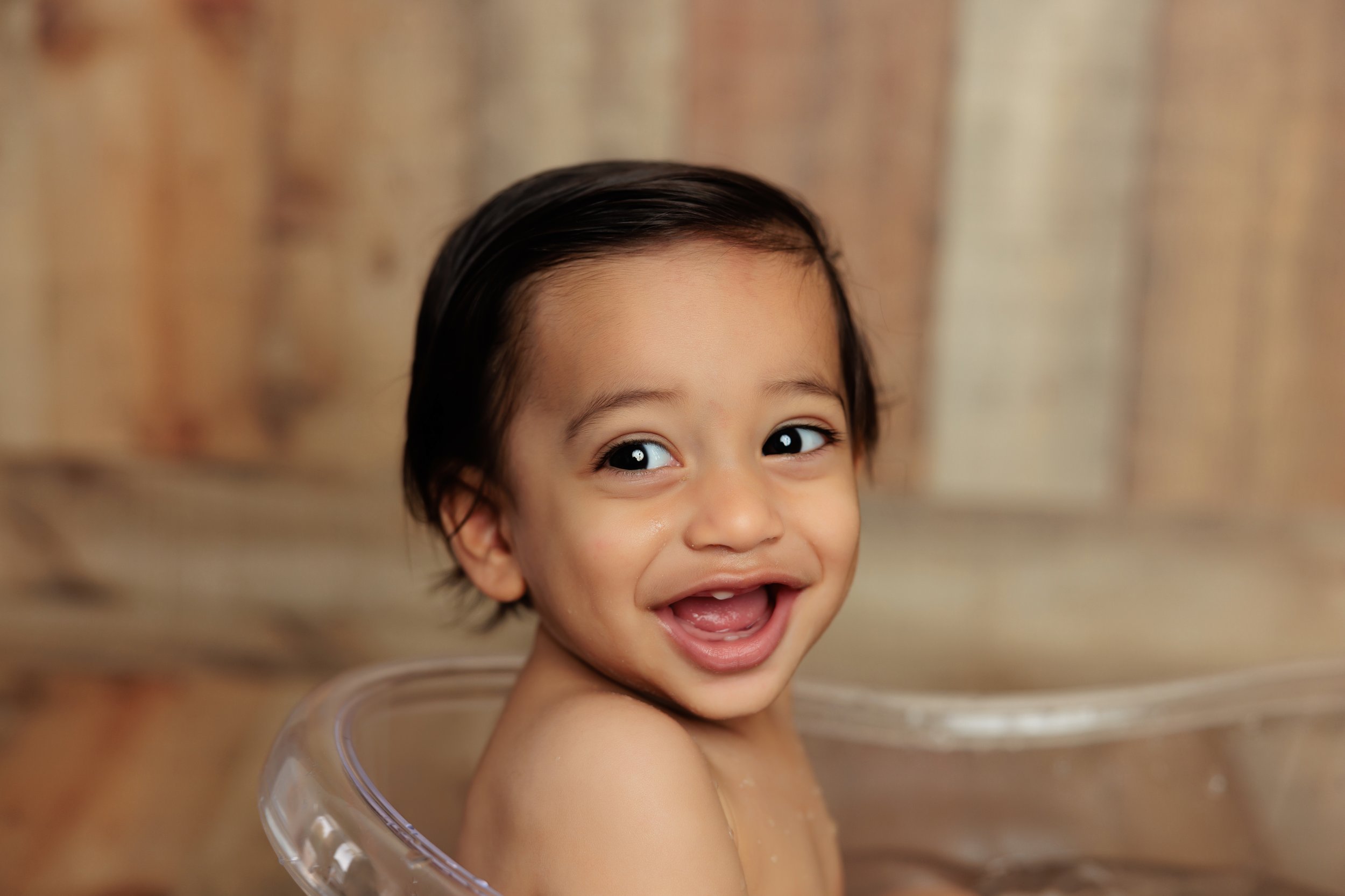 Smiling young child with dark hair taking a bath in a clear plastic tub, wooden background.