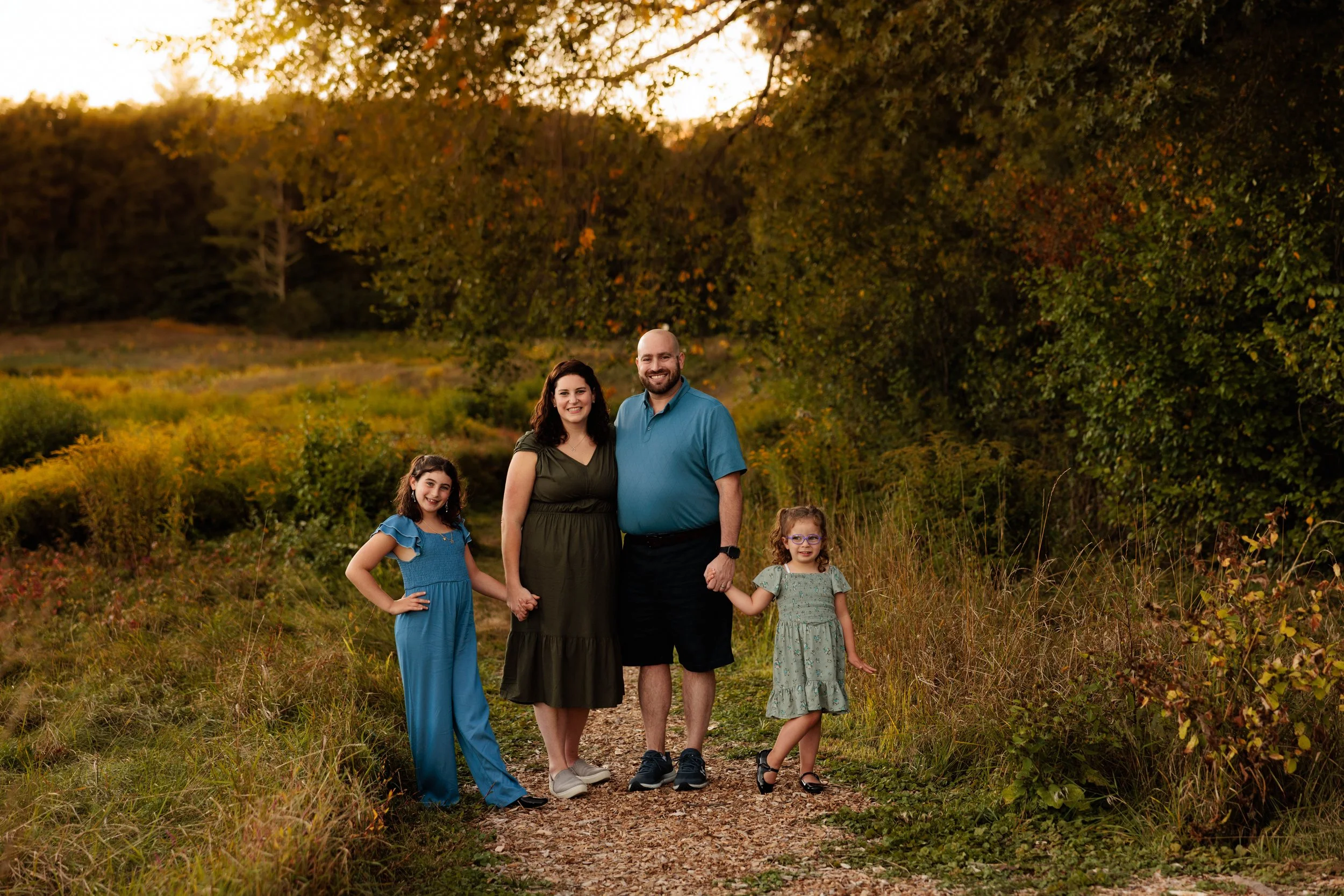 Family of four standing on a dirt path in a lush, green park during sunset. The mother, father, and two young daughters are smiling, holding hands, and dressed in casual summer clothes.