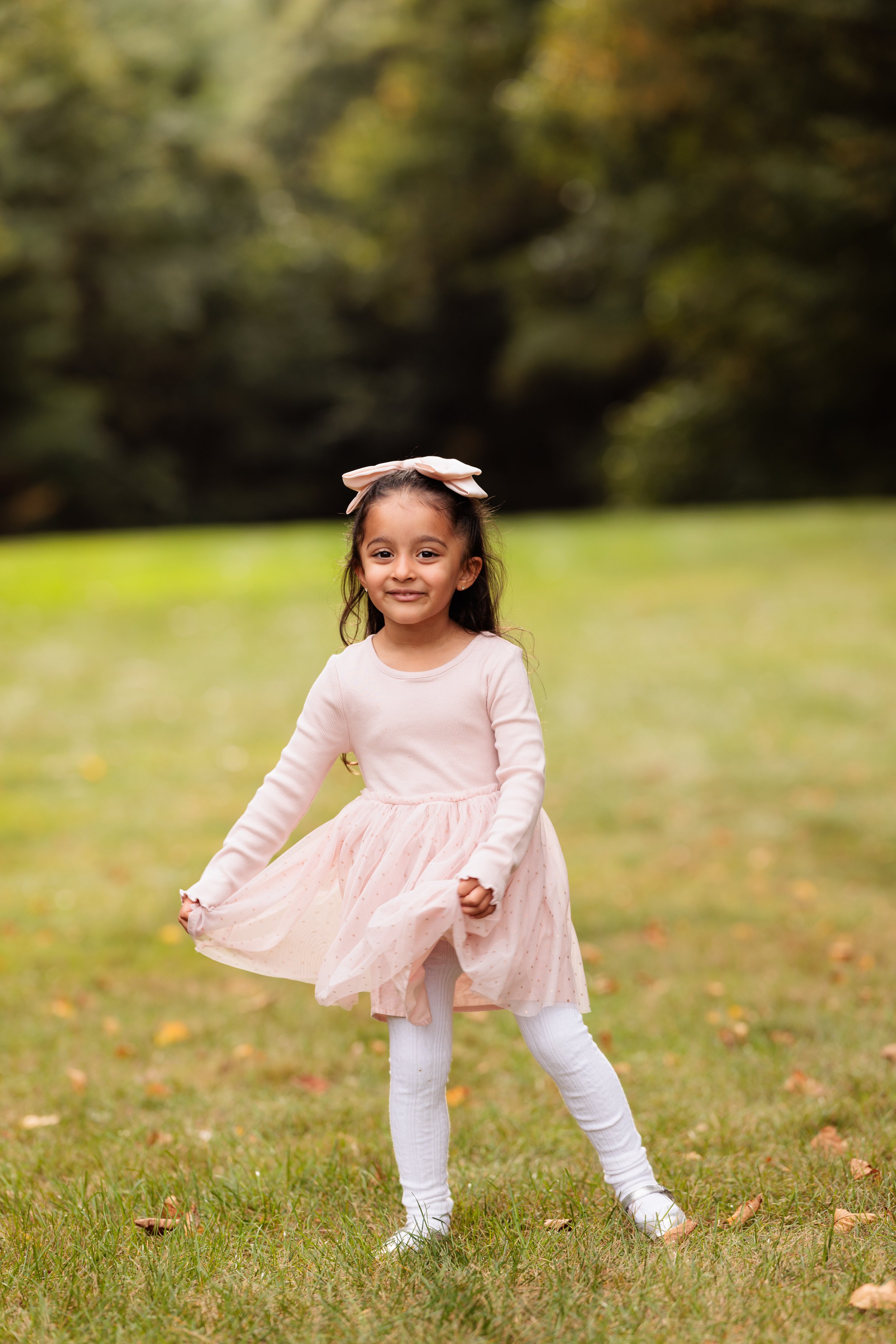A young girl in a pink dress with a bow in her hair standing on grass outdoors, smiling at the camera.