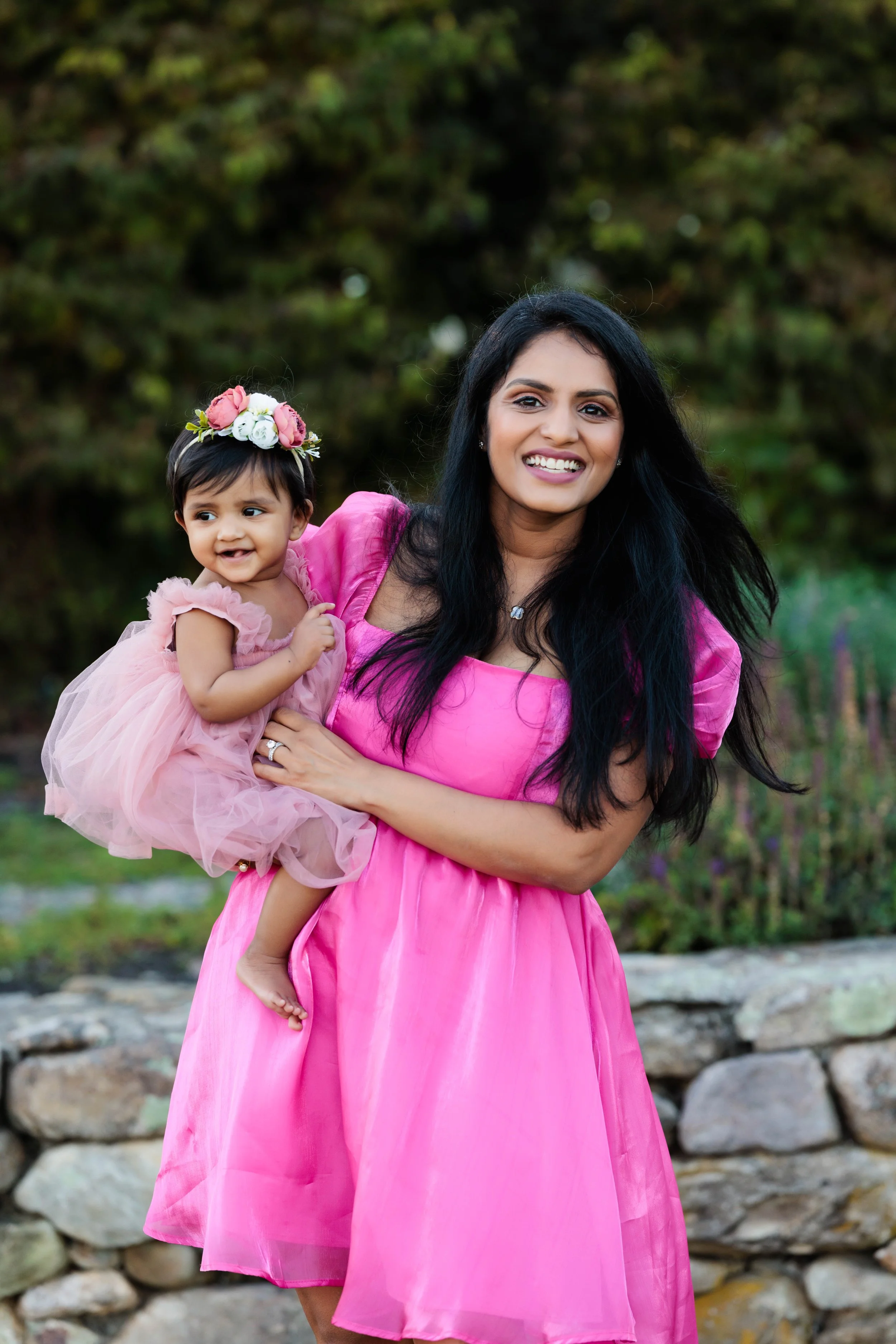A woman in a pink dress holding a smiling young girl in a pink dress and flower crown outdoors.