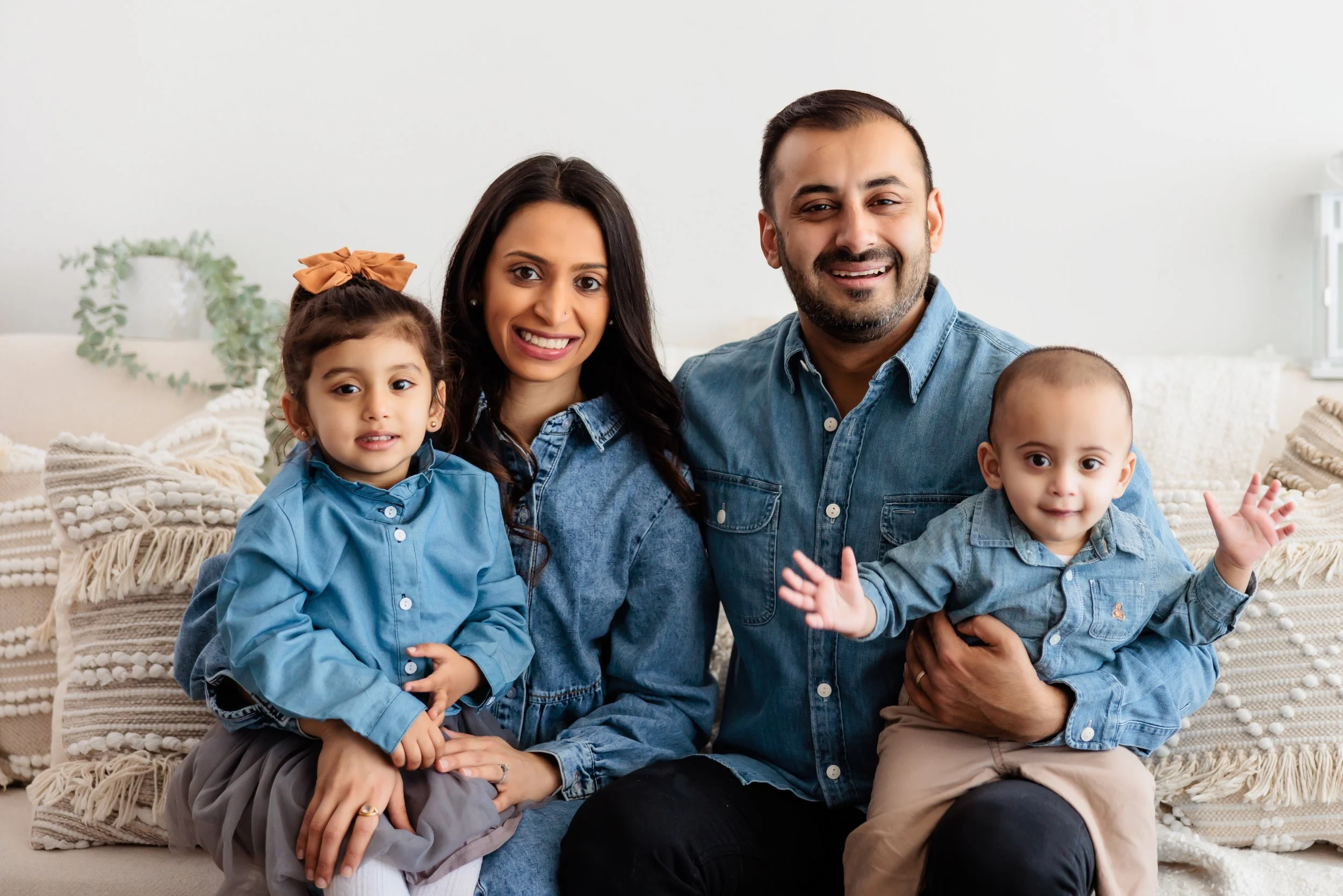 Family of four sitting on a couch, smiling, with a white wall and decorative pillows in the background.