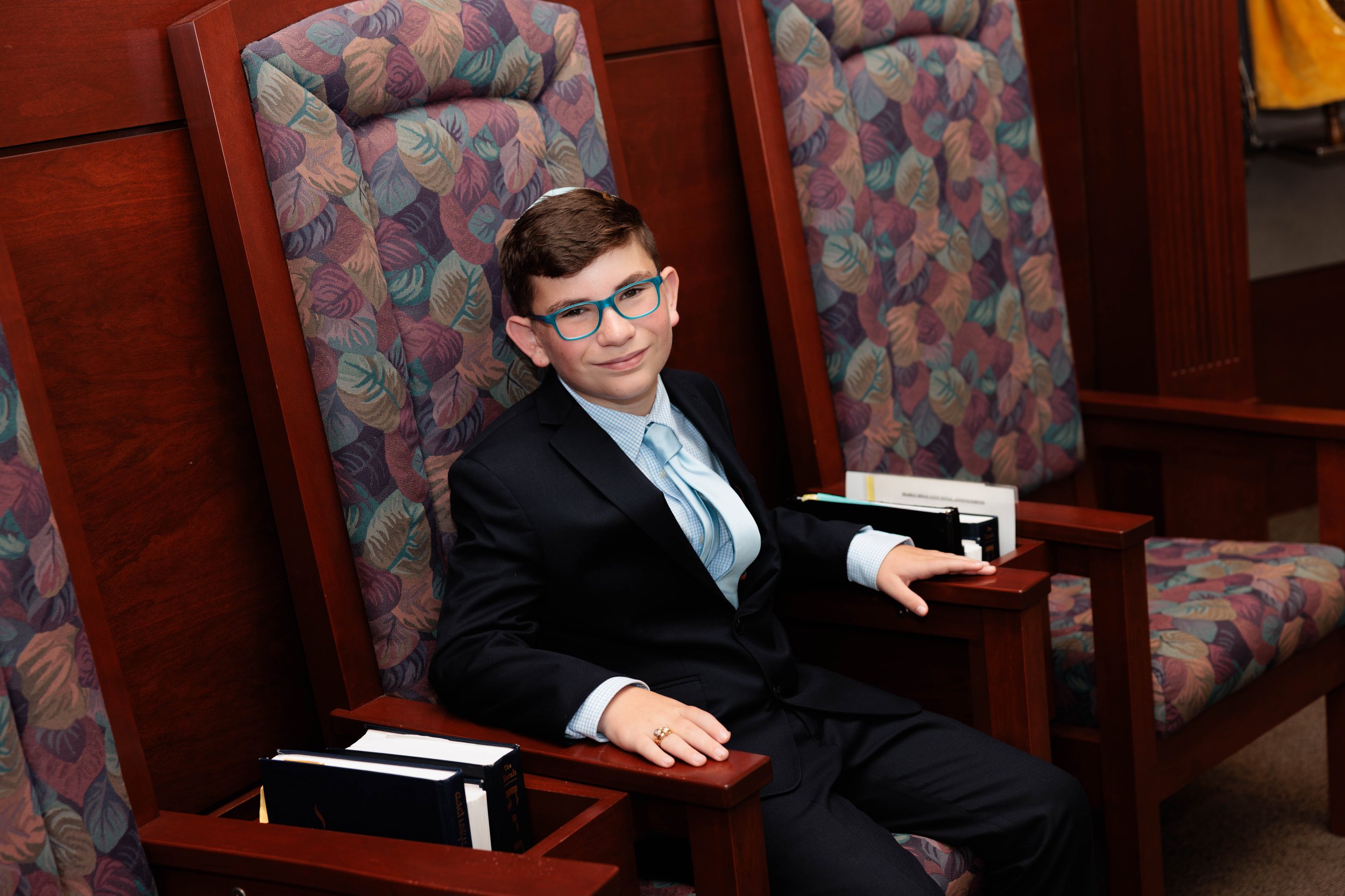 A young boy with brown hair and glasses, dressed in a black suit, white shirt, and light blue tie, sitting in a wooden courtroom chair with patterned fabric, surrounded by similar chairs with books on the arms.