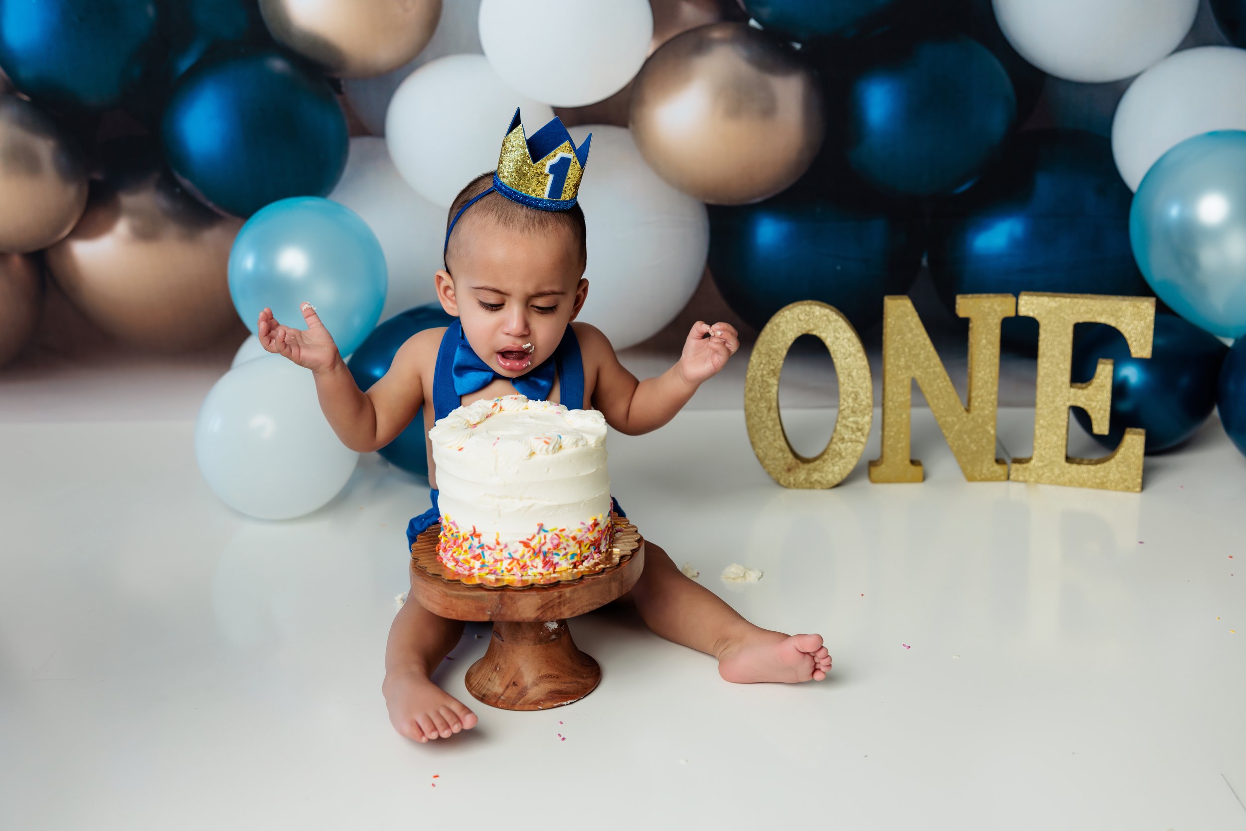 A young child wearing a birthday hat, blue bow tie, and blue outfit, sitting on the floor with a partially eaten birthday cake in front of them. The child appears to be upset or surprised. In the background, there is a balloon arrangement in shades o