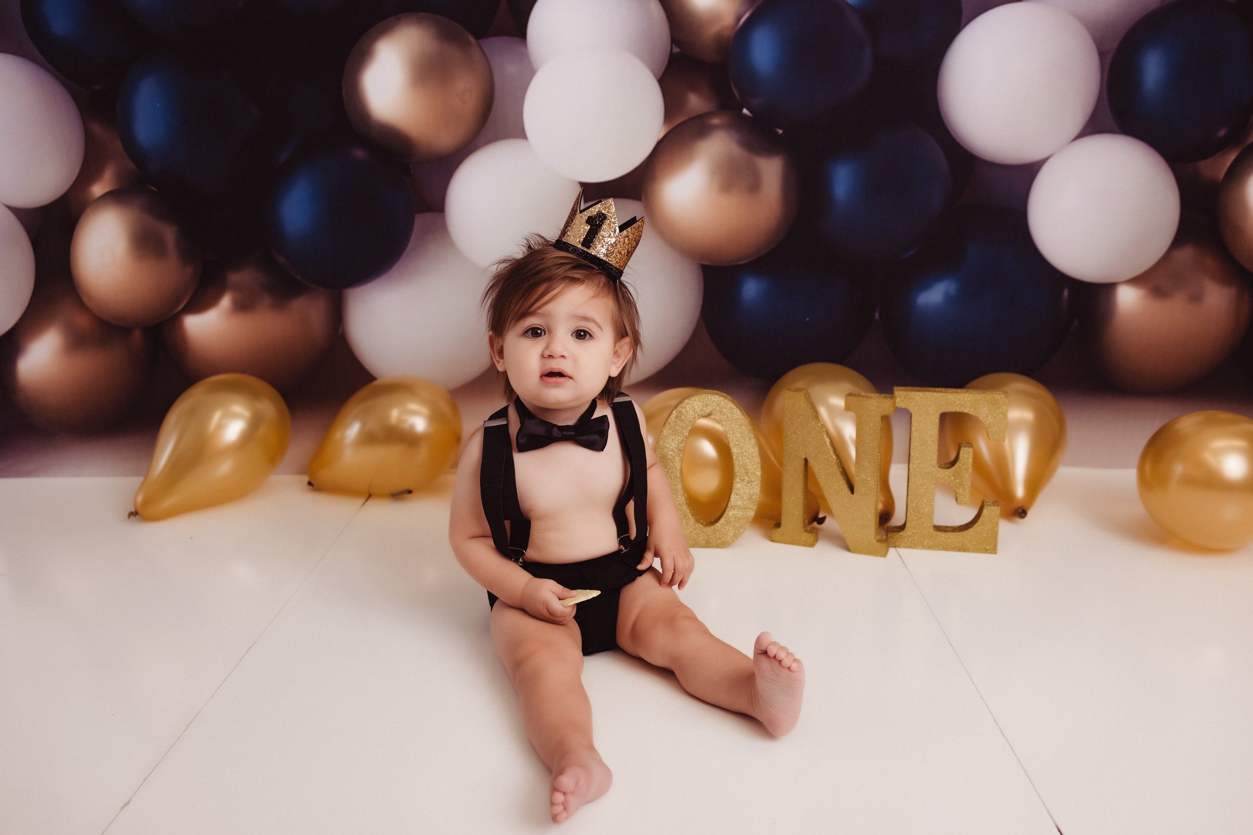 A toddler sitting on the floor in front of a backdrop of gold, white, navy, and bronze balloons, with large gold letter blocks spelling 'ONE' and a gold crown headband on his head for a first birthday celebration.