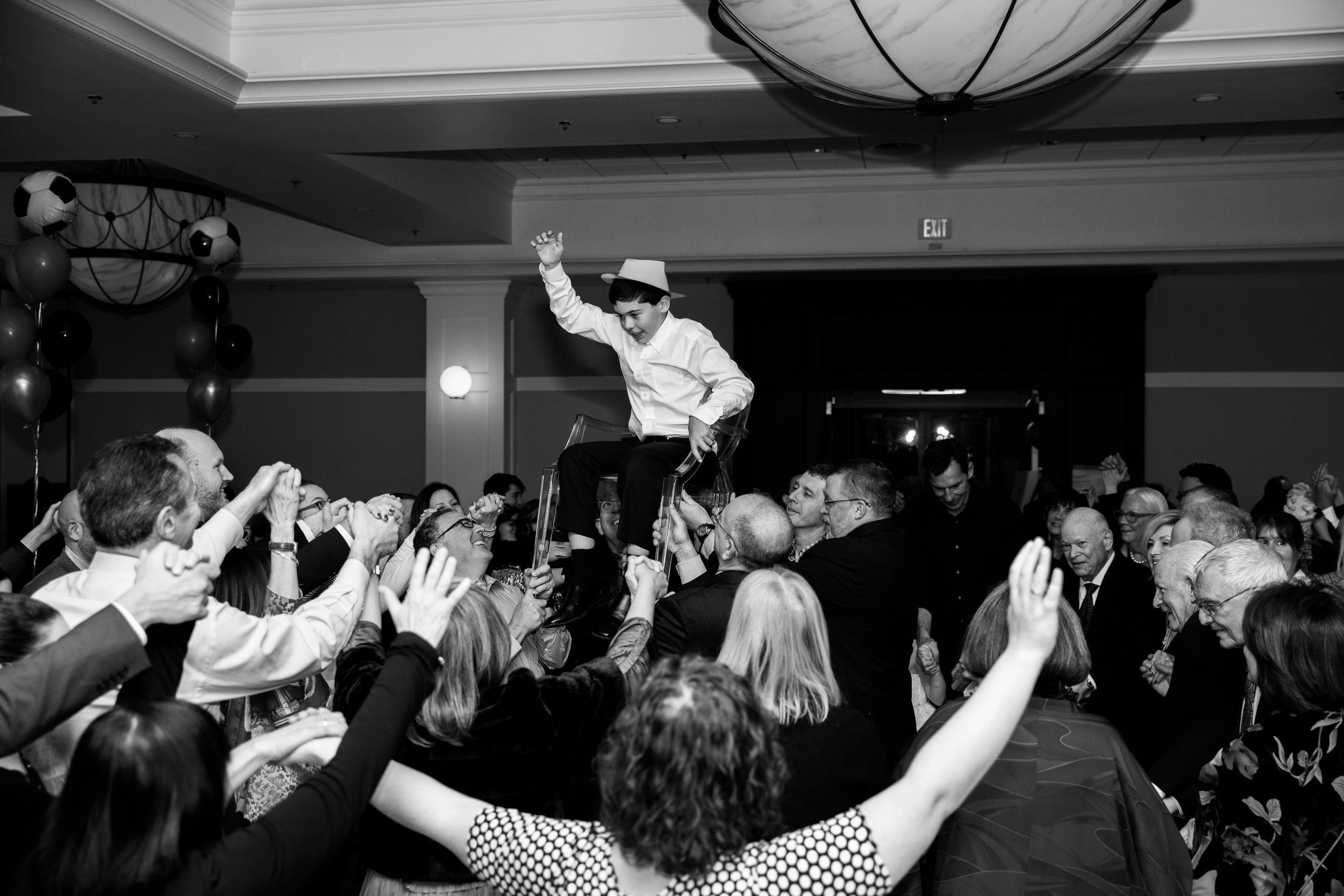 A boy in a white shirt and a paper hat sits on a chair held up by adults at a celebration or party, surrounded by a crowd of smiling and clapping people.