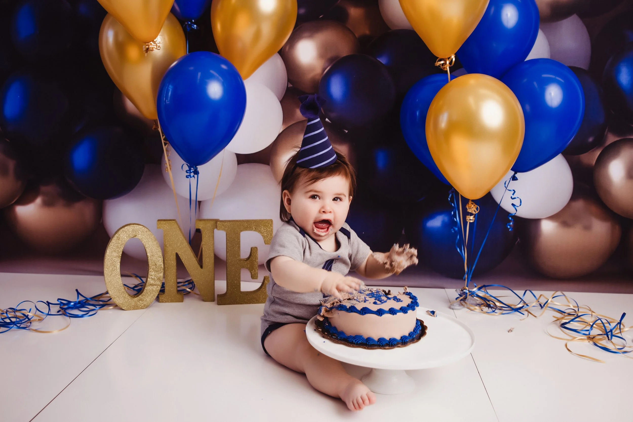 A young child celebrating a first birthday with a cake, wearing a striped birthday hat, and sitting on a white surface in front of a backdrop of blue, gold, black, and white balloons with a gold 'ONE' sign.