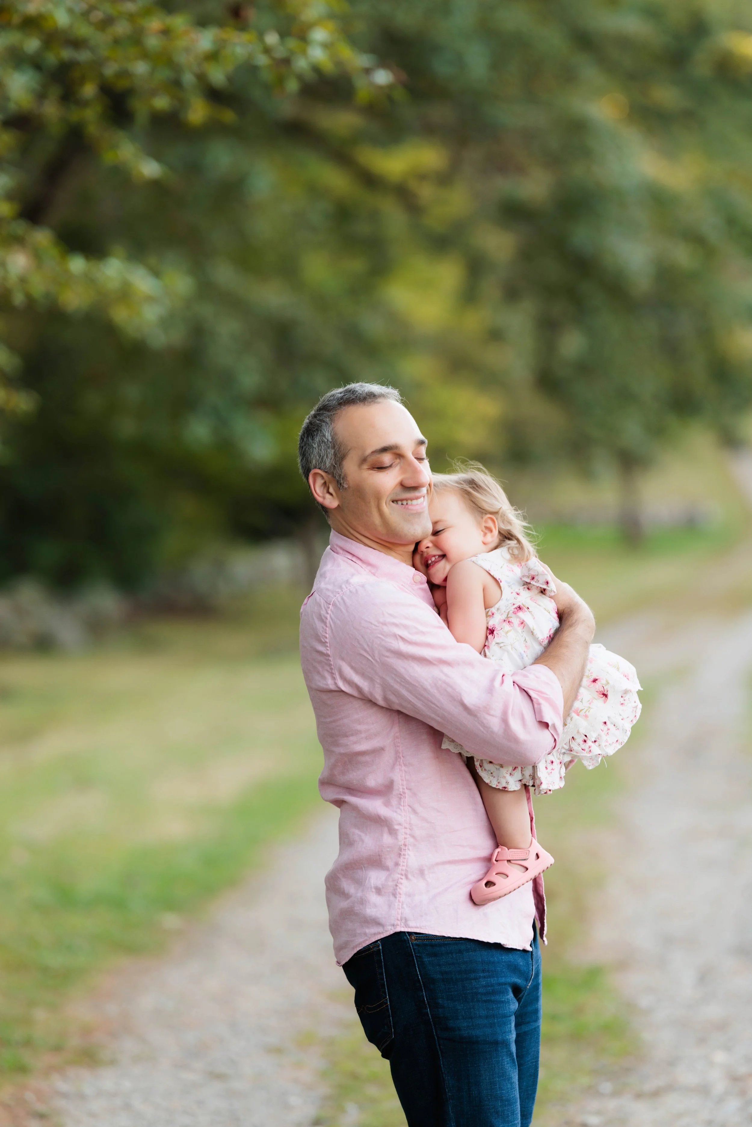 A man holding a young girl in a pink dress and shoes, outdoors on a grassy and gravel path with trees in the background.