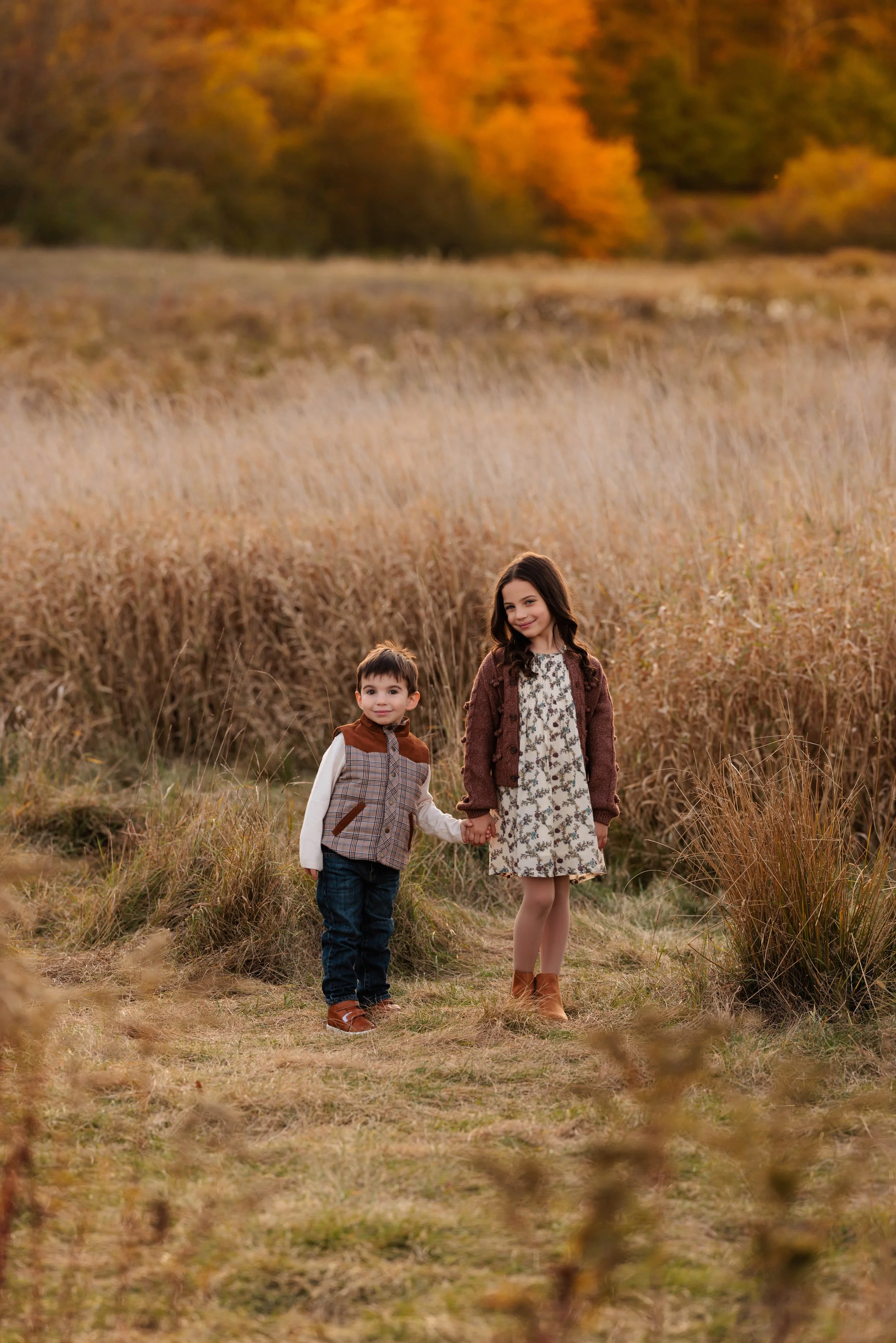 A young girl and boy holding hands walking through a grassy field during fall, with trees displaying orange and yellow foliage in the background.