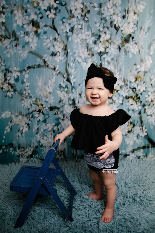 A smiling toddler girl with a black bow headband, wearing a black top and patterned shorts, standing on a fuzzy teal rug next to a small blue wooden bench with a floral backdrop.