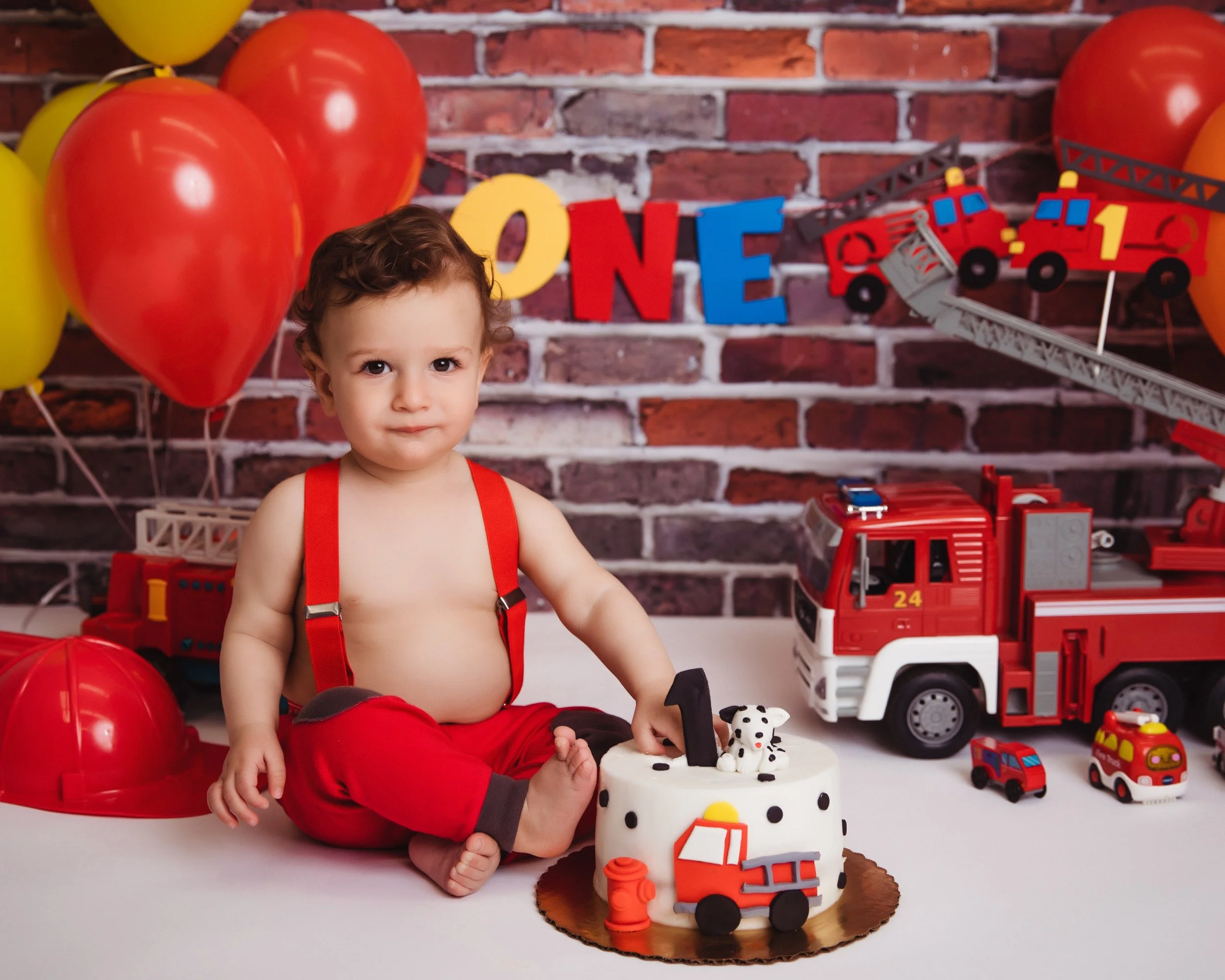 A young child with curly hair and a serious expression, sitting on the floor at a birthday party. The child is wearing red suspenders and red pants, with one hand on a firefighter-themed birthday cake. Behind the child are red and yellow balloons, a 