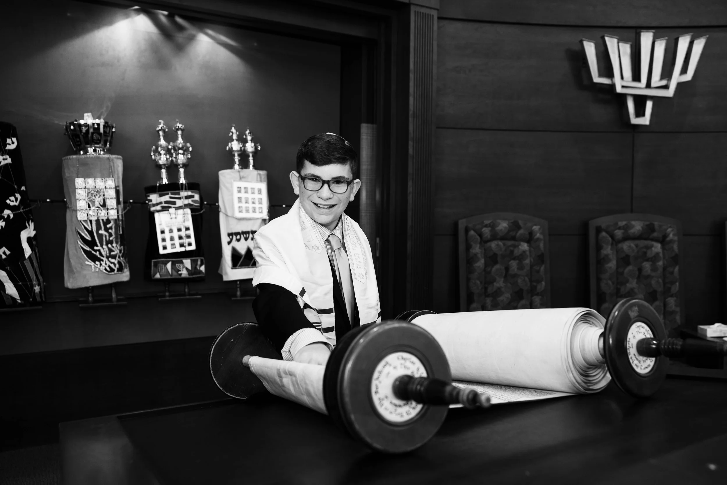 A young man wearing glasses, a tie, and a tallit, smiling and rolling out a Torah scroll on a table in a room decorated with Jewish symbols and artifacts.