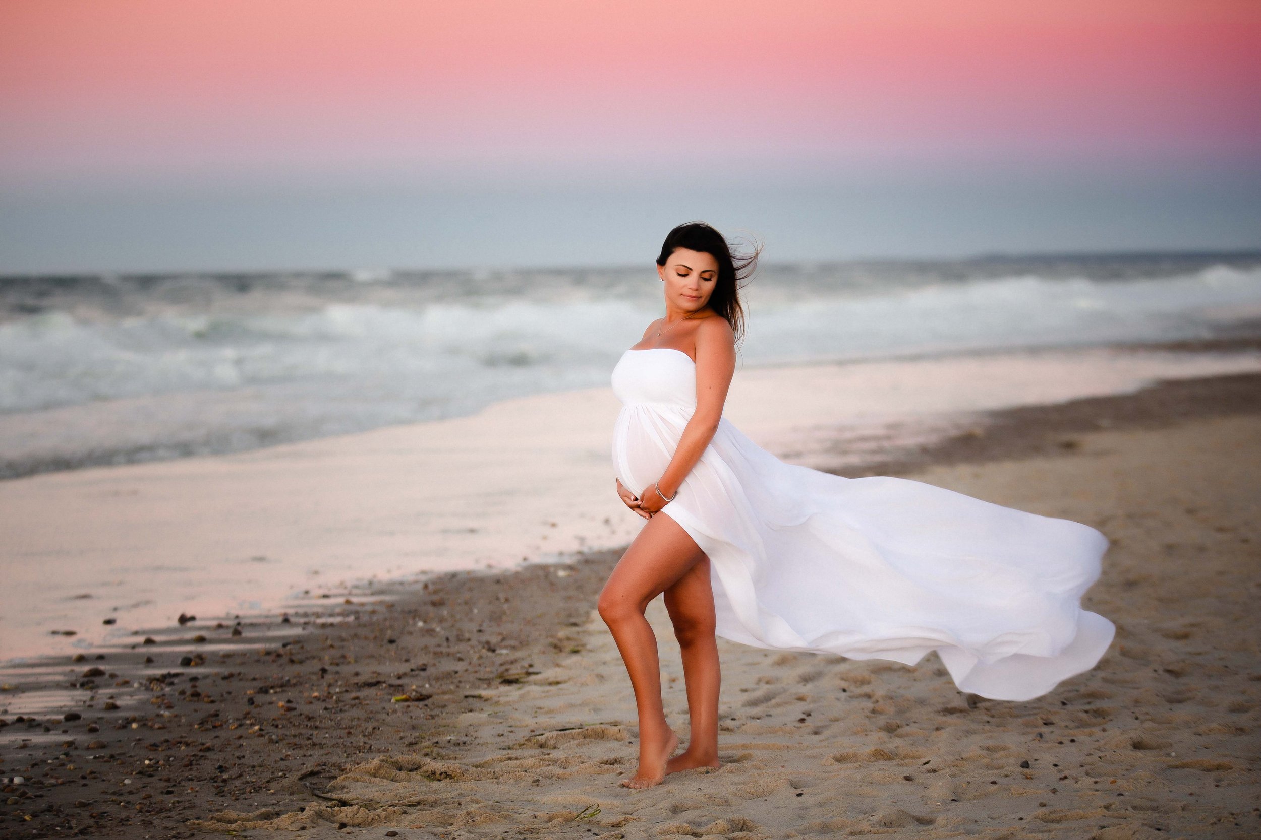 Pregnant woman in a white dress standing barefoot on the beach with the ocean and pink sunset sky in the background.
