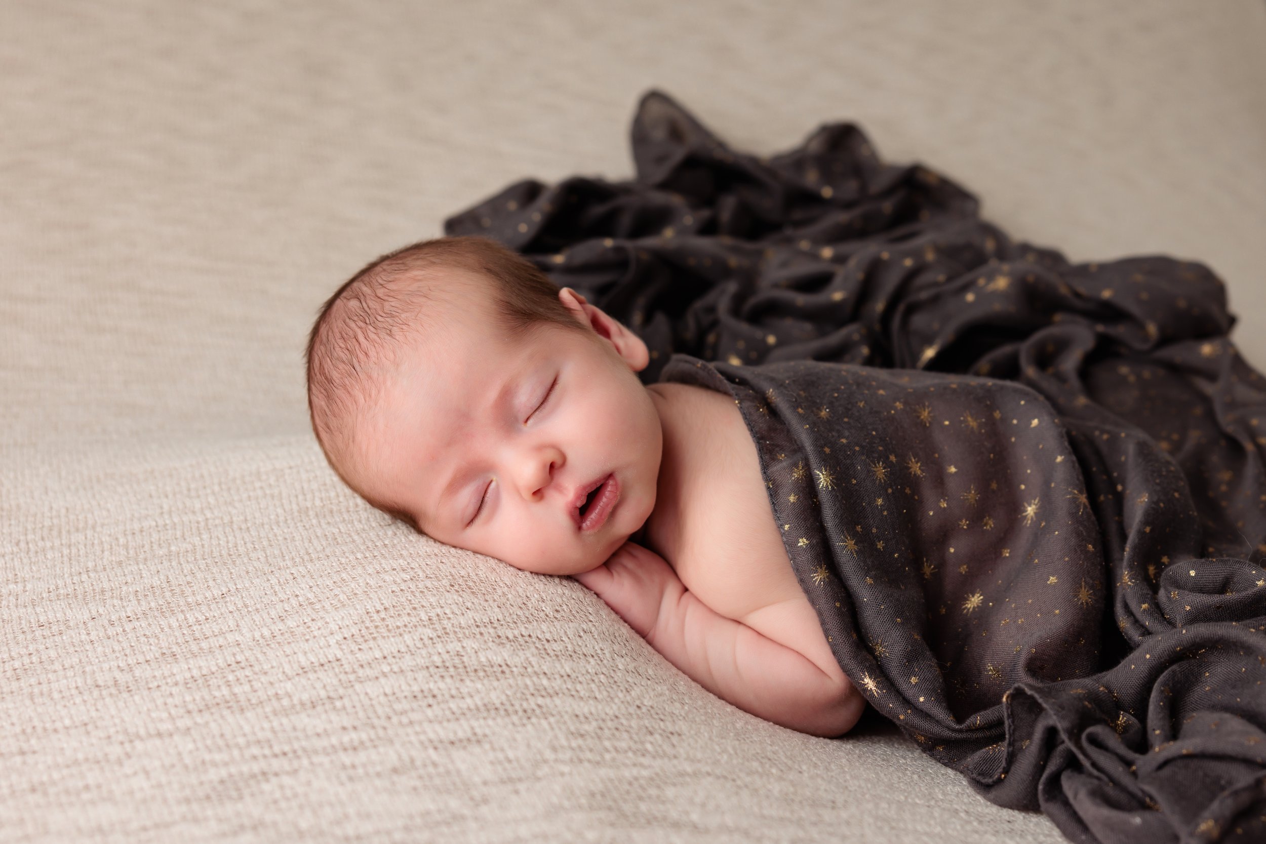 A young baby sleeping peacefully on a beige textured surface, partially covered with a dark blanket with gold star patterns.