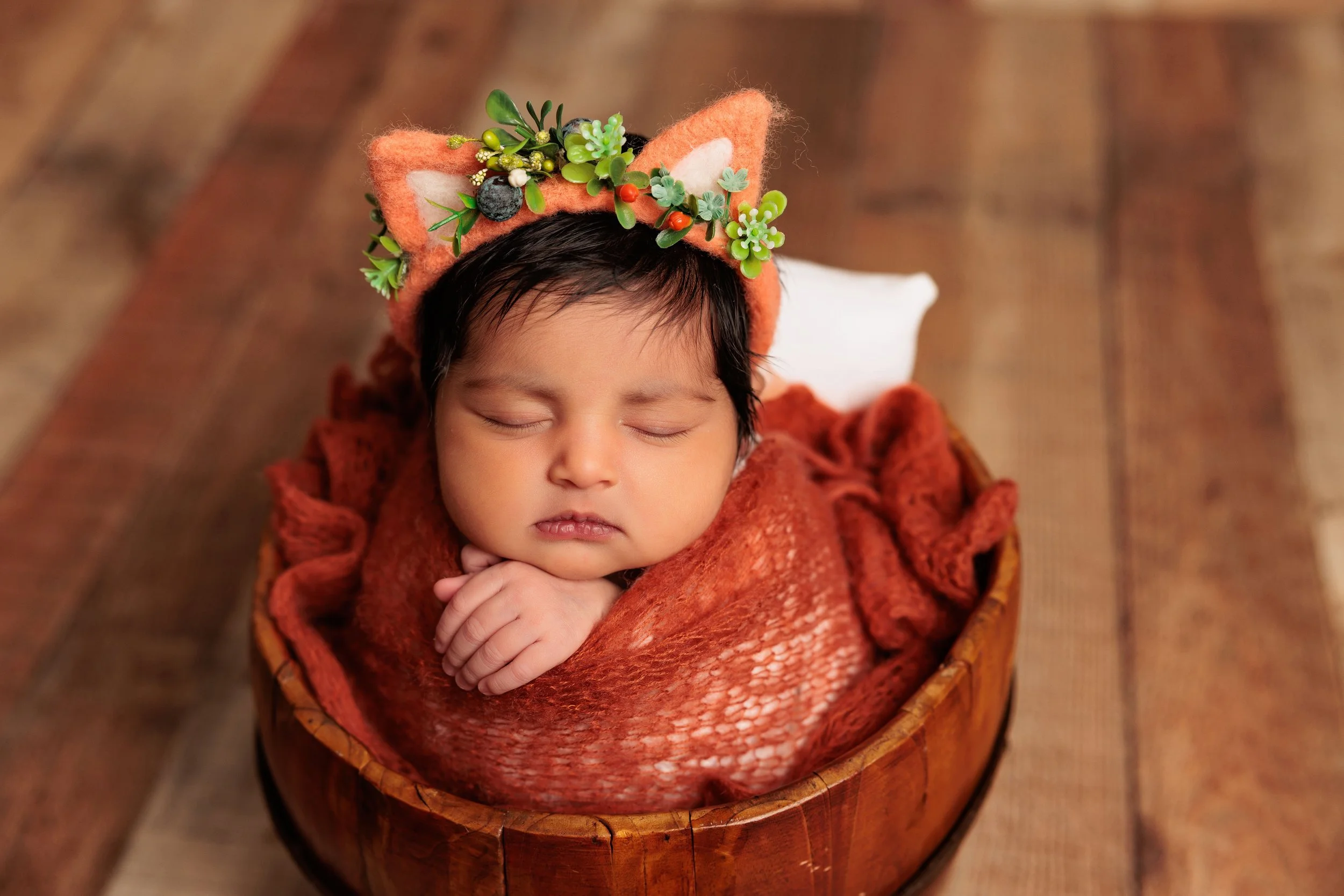 A sleeping baby with dark hair, wrapped in a reddish-brown blanket, lying inside a wooden bowl on a wooden floor. The baby wears a fuzzy orange headband with green flowers and leaves, and has its hands tucked under its chin.
