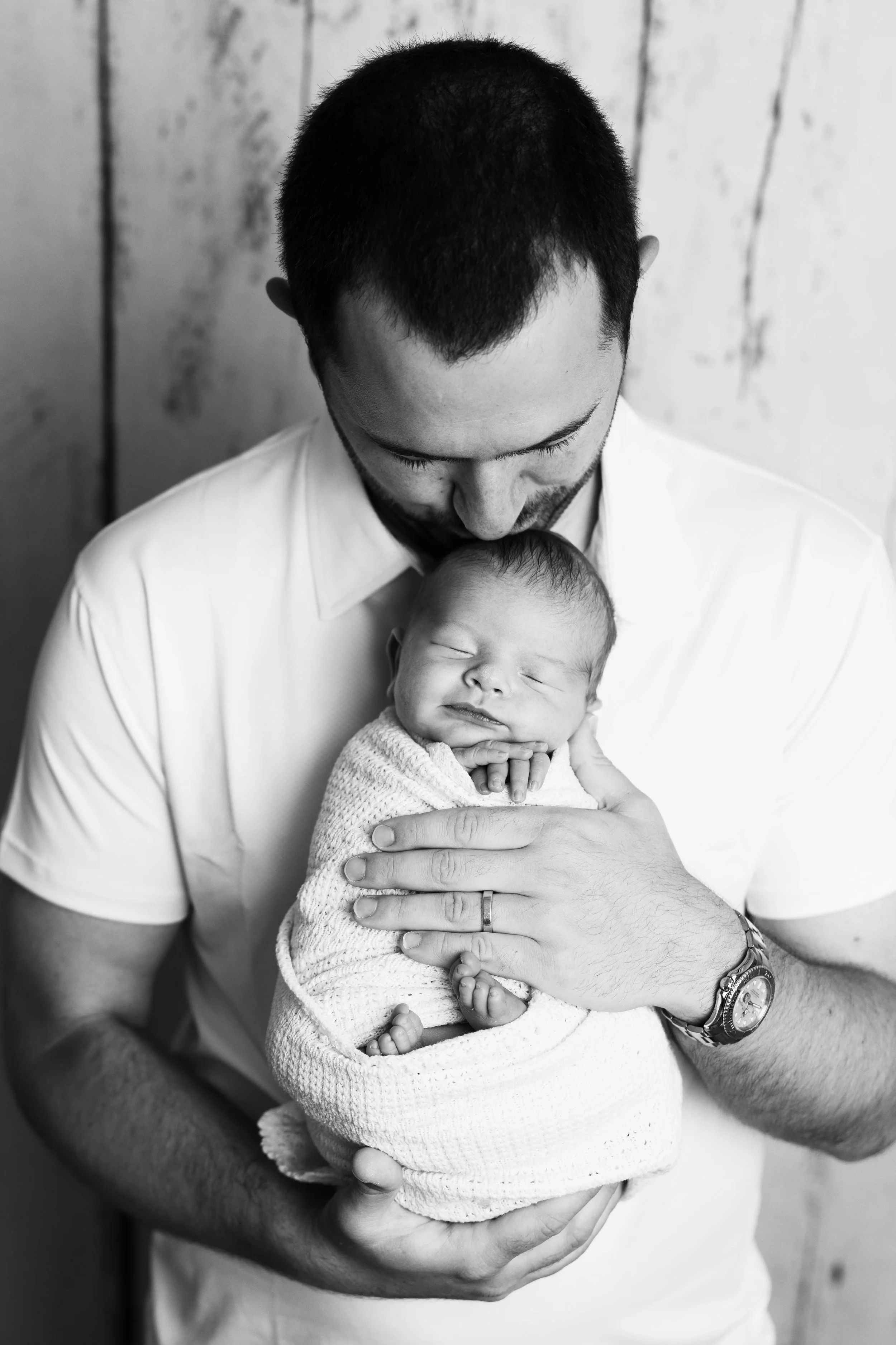 A man holding a sleeping newborn baby wrapped in a blanket, with the man gently kissing the baby's head, in a close-up black and white photo.