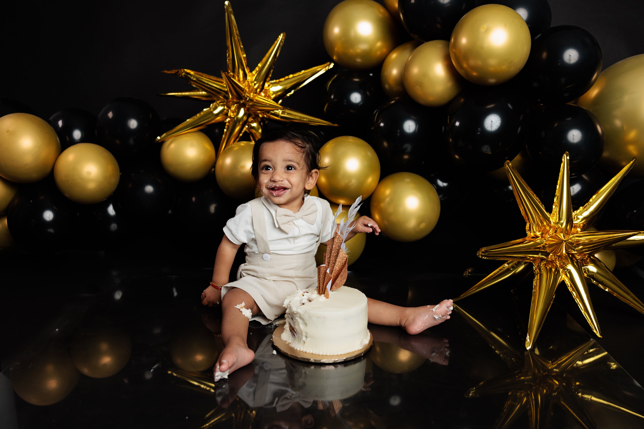 A young smiling child sitting on a black reflective surface surrounded by black and gold balloons and golden star-shaped balloons behind her. She is celebrating her birthday with a small white cake decorated with feathers and copper stars. The child 
