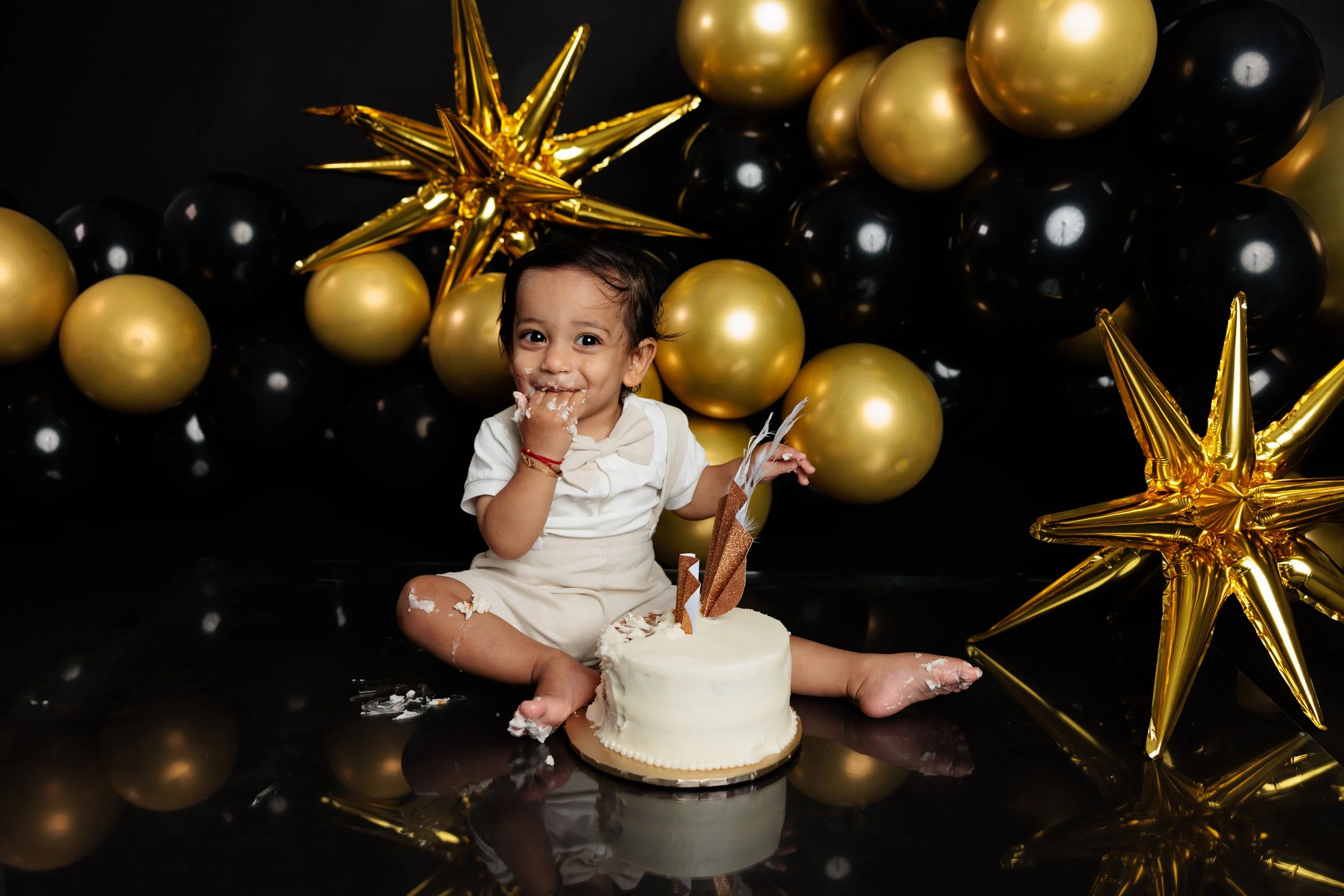 A young child celebrating a birthday, sitting on a black reflective surface with a cake in front. The child is smiling with cake and frosting on their face, surrounded by black and gold balloons and shiny star-shaped decorations.