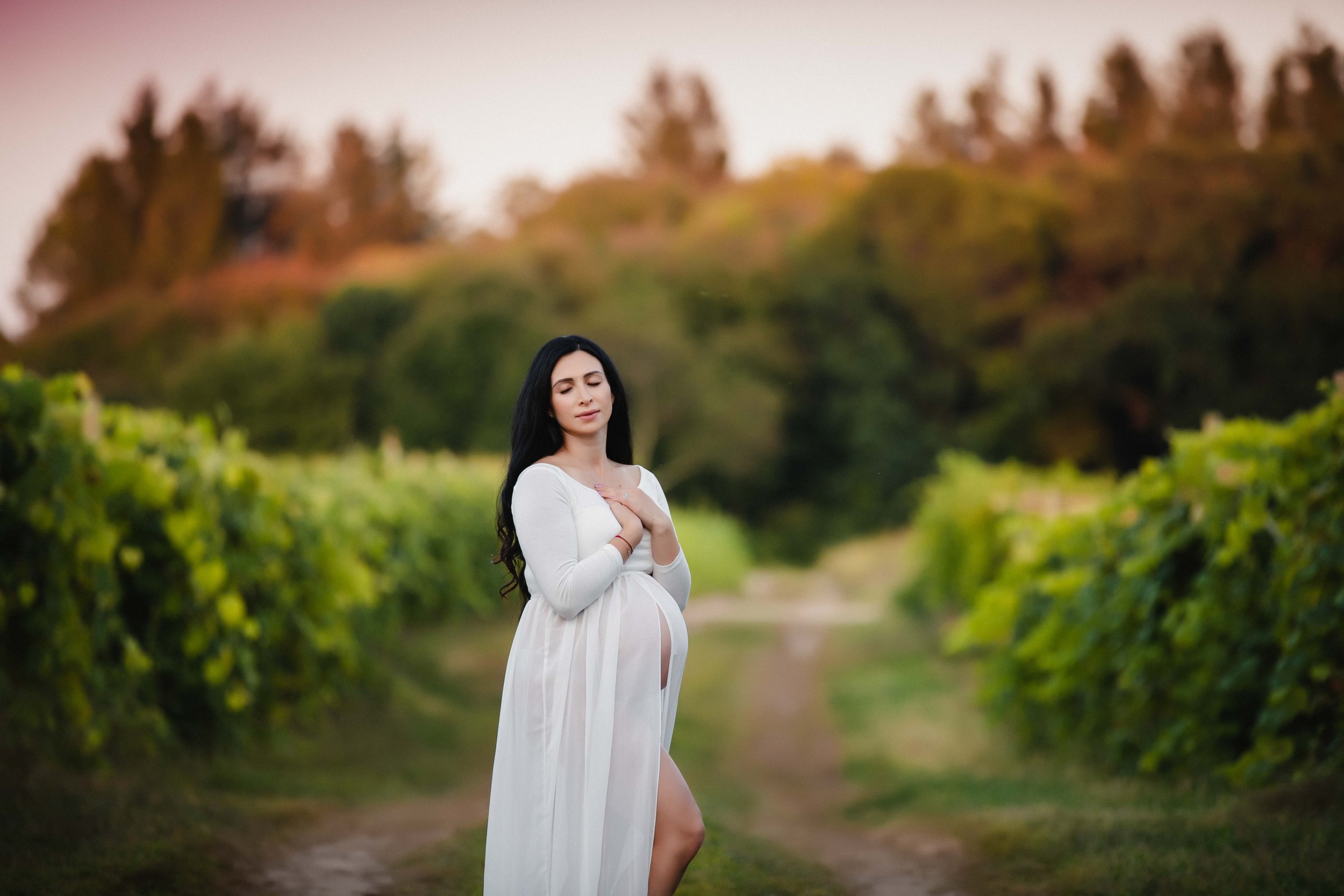 A pregnant woman in a white dress standing on a dirt path amid green vineyard rows, with a blurred hillside background at sunset, holding her hands over her chest with a serene expression.