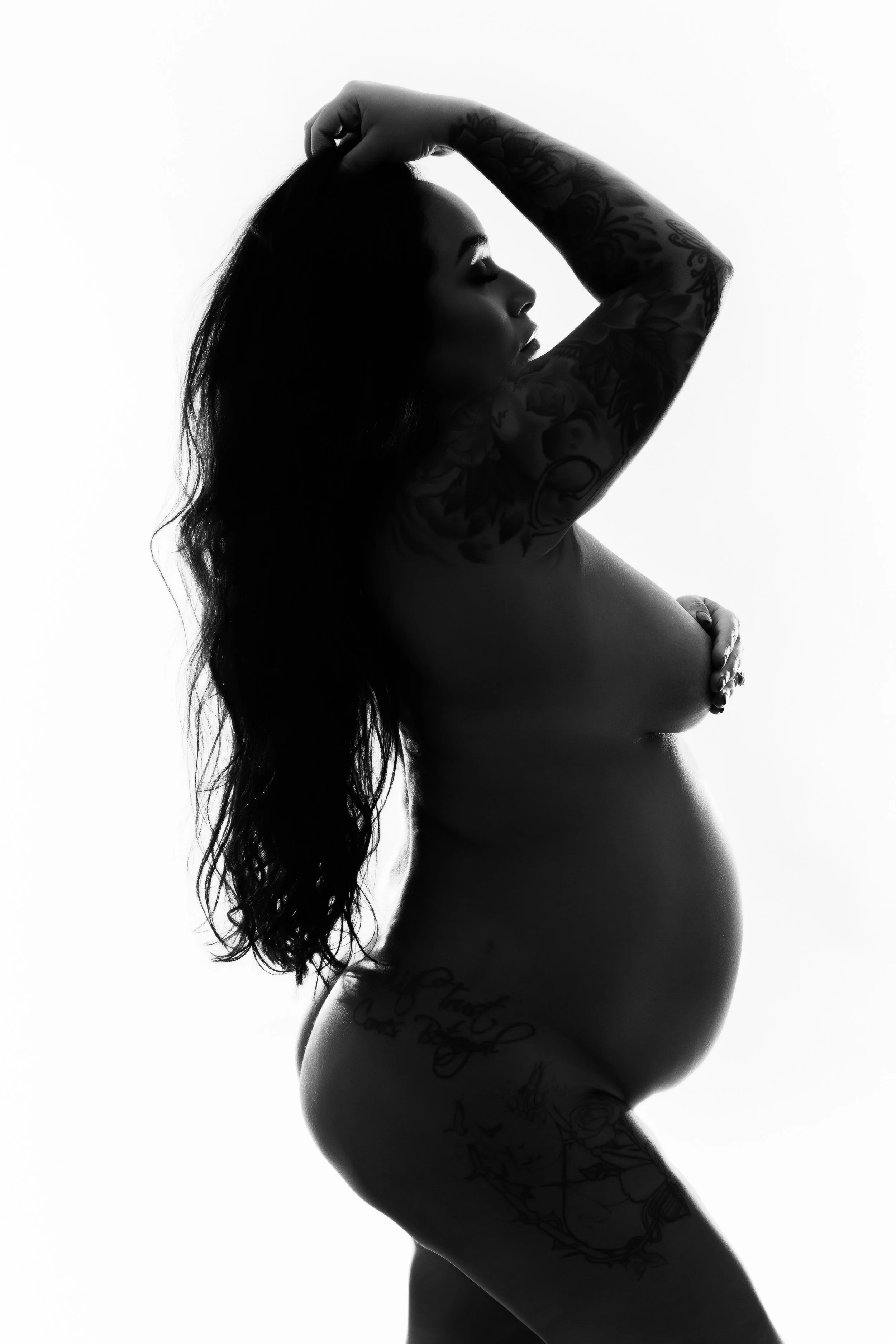 Silhouette of a pregnant woman with long dark hair, tattoos on arms and thighs, holding her belly with one hand and resting the other on her head, against a white background.