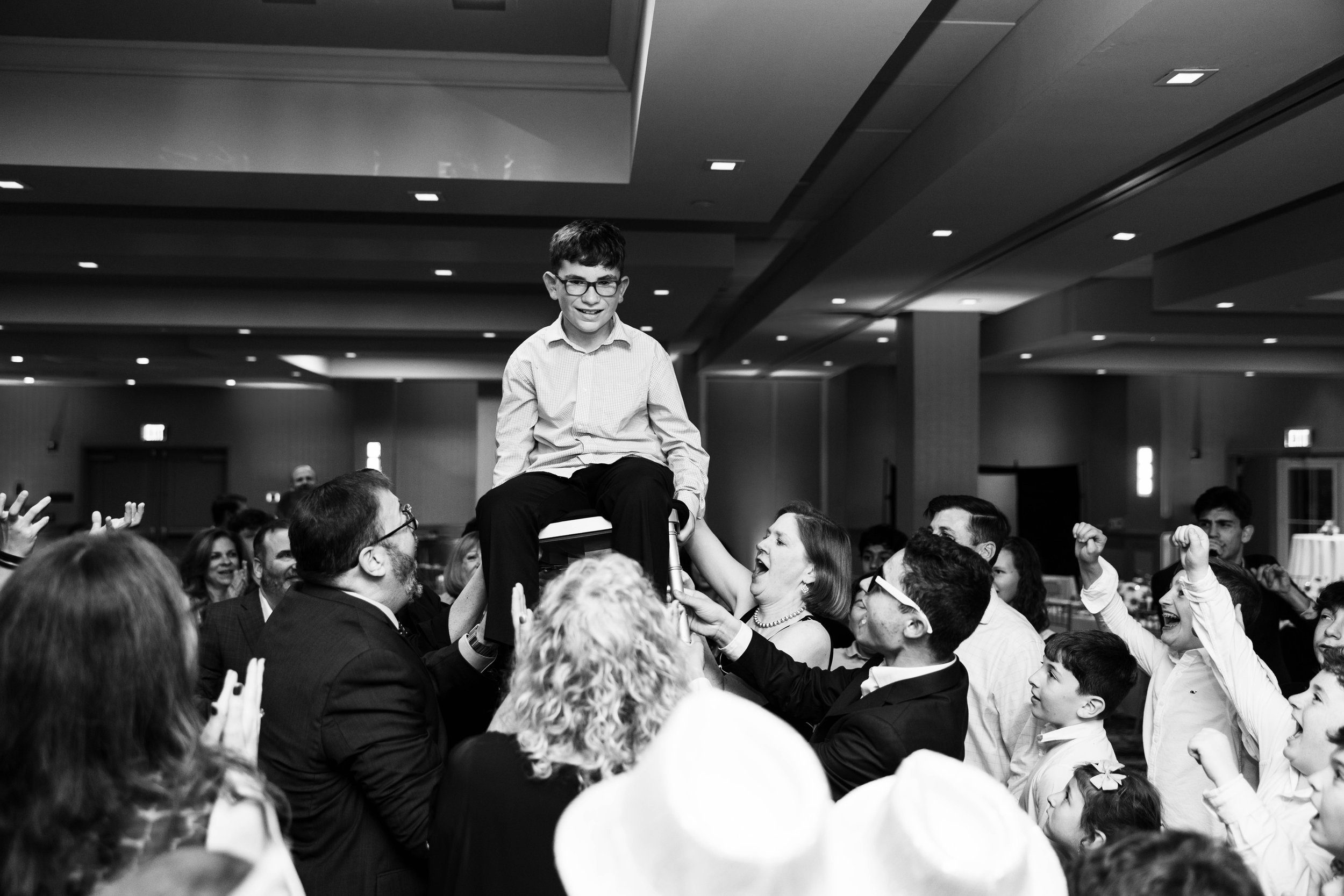 A boy sitting on a chair being lifted by a group of people, smiling and celebrating at a social event or celebration in a large decorated hall.