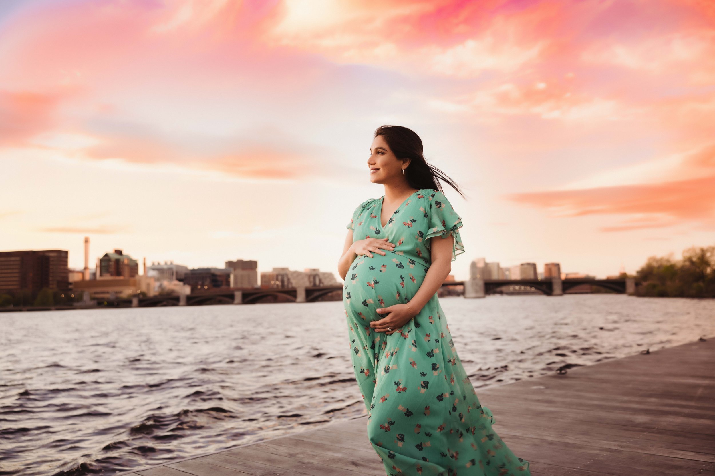 A pregnant woman in a green floral dress standing on a riverside promenade during sunset, smiling and holding her belly.