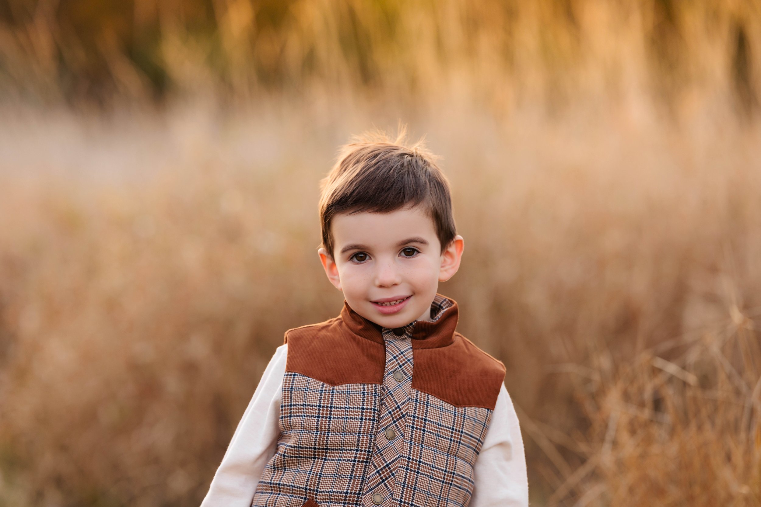 A young boy with dark hair and brown eyes standing outdoors in a field of tall, golden grass during sunset, wearing a brown and plaid vest over a white shirt.