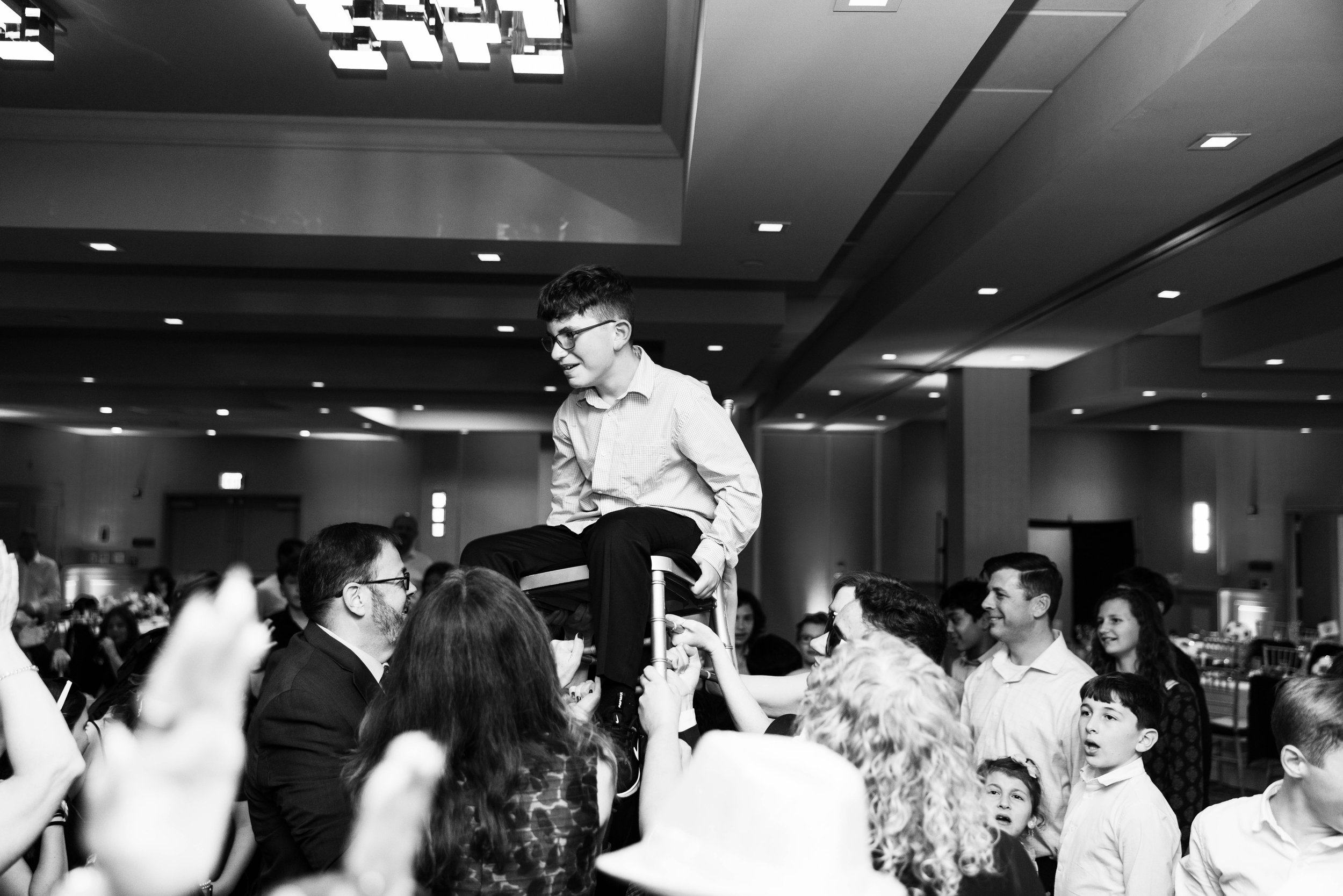 A person in a dress shirt and glasses is seated on a chair held up by multiple people in a crowded, indoor event space or banquet hall, with several onlookers and participants around.
