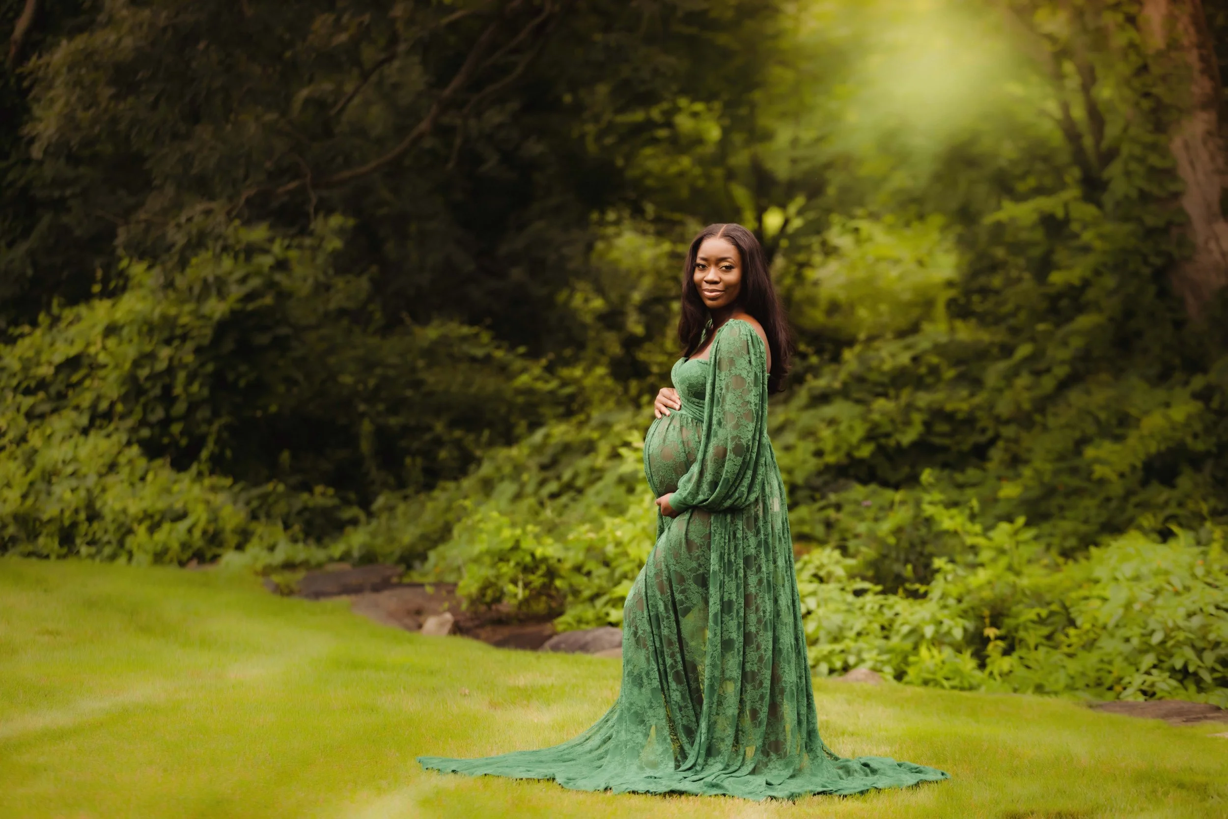 A pregnant woman in a flowing green lace dress standing on grass in a lush, green outdoor setting.