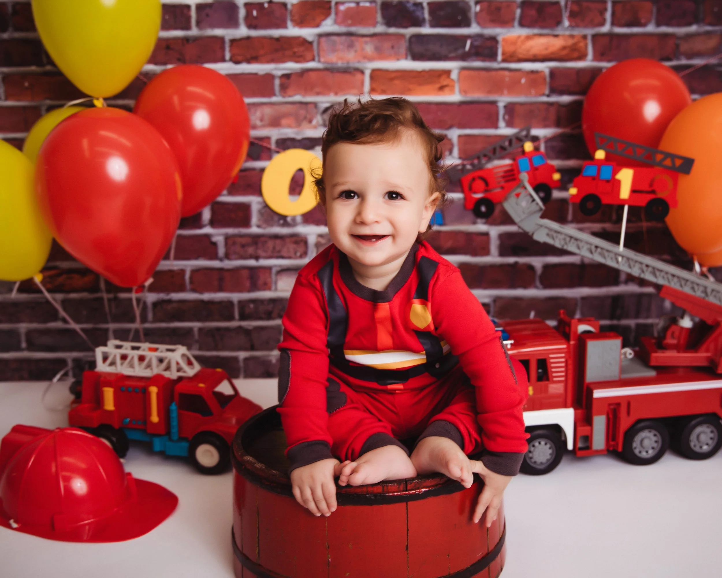 A smiling young child dressed in a red firefighter costume sitting on a red barrel surrounded by toy fire trucks and red and yellow balloons against a brick wall background.