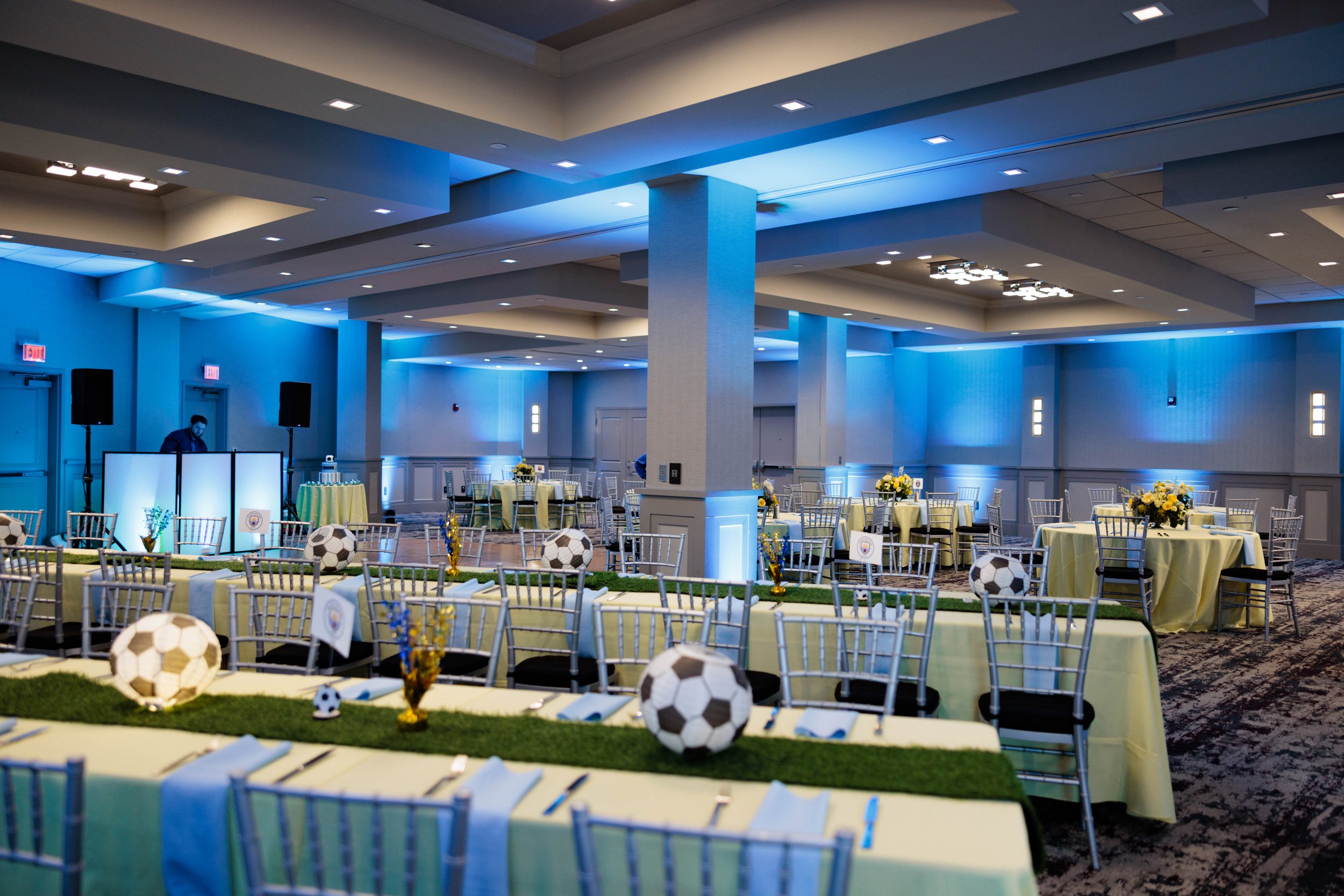 Event space decorated with soccer-themed centerpieces, featuring paper soccer balls on tables, yellow flowers, and blue lighting along the walls, set up for a celebration or gathering.