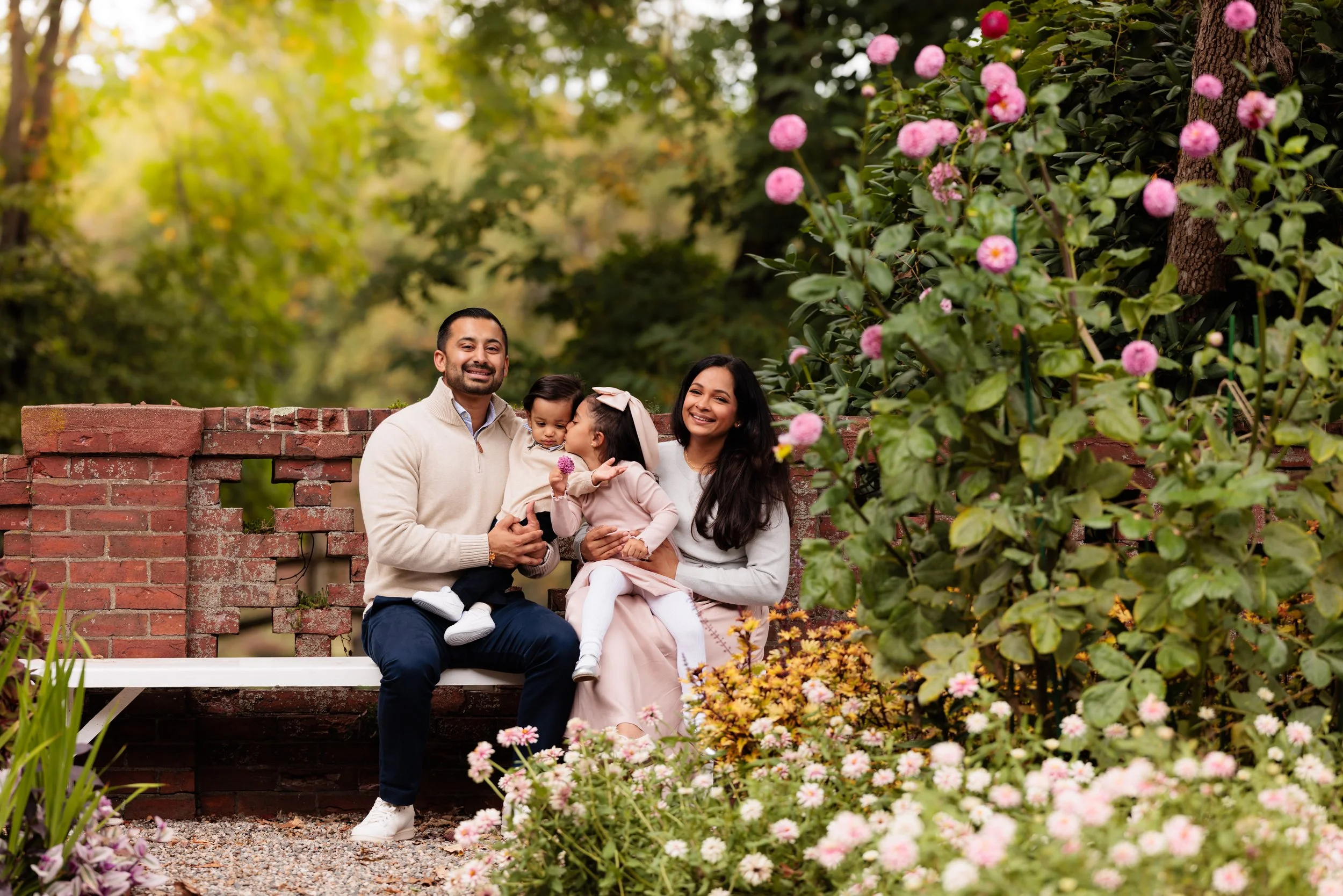 A family of four sitting on a white bench in a garden with blooming pink flowers and green trees in the background. The man and woman are smiling, with the woman holding a young girl and the man holding a toddler.