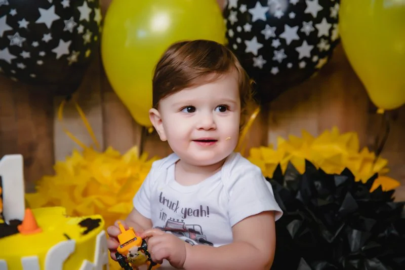 A young child with brown hair sitting at a birthday party with yellow and black balloons and decorations.