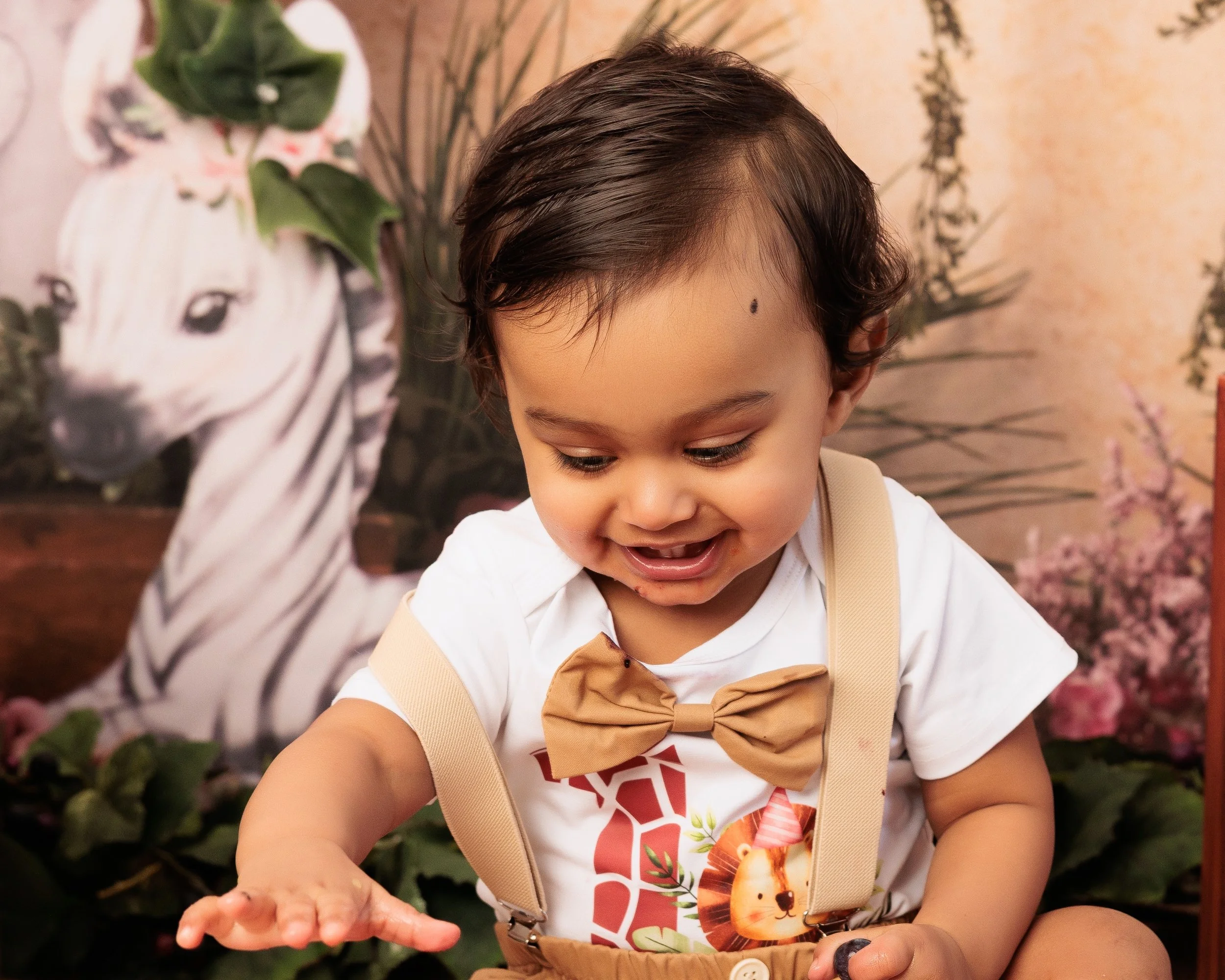 A young child with dark hair smiling and playing in front of a zebra-themed backdrop with green plants and pink flowers.