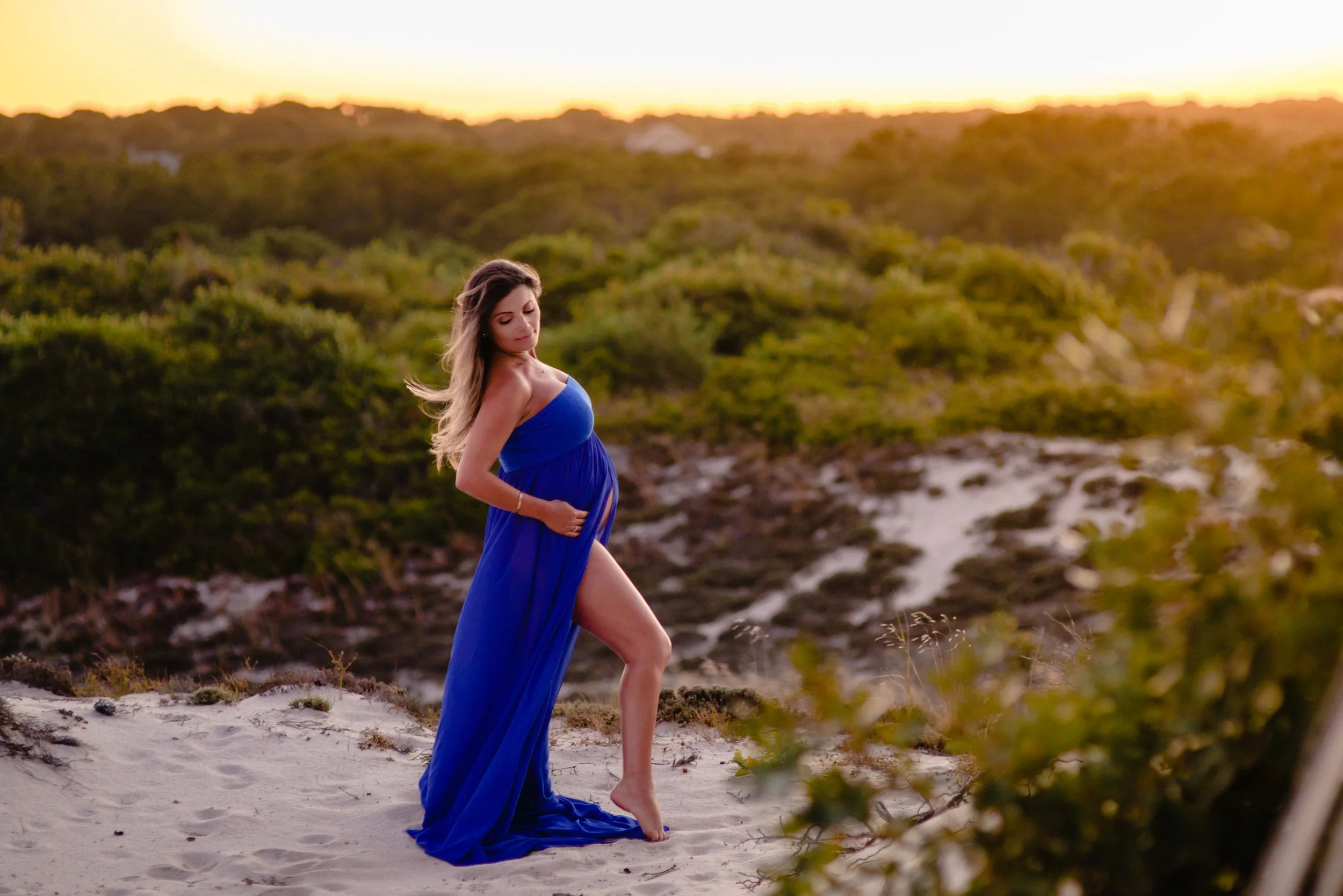 A pregnant woman in a blue dress standing barefoot on sand dunes during sunset, with green bushes and a yellow-orange sky in the background.