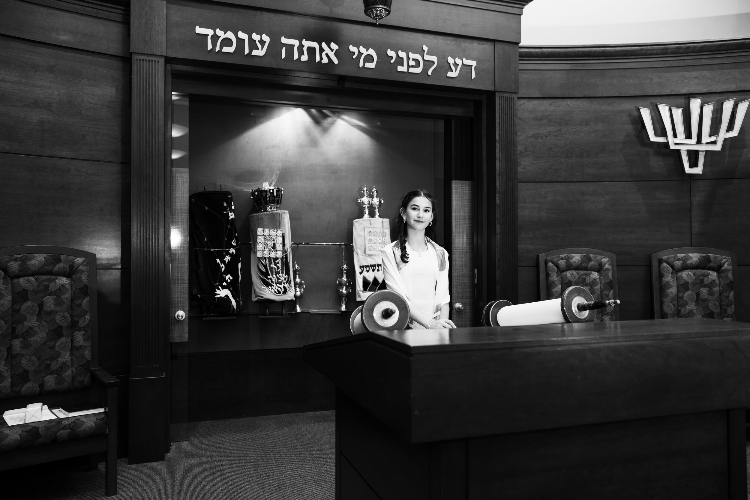 A young girl standing behind a wooden table in a synagogue, with menorahs and Torah scrolls displayed in the background, Hebrew text on a sign above her, and a menorah symbol on the wall to the right.