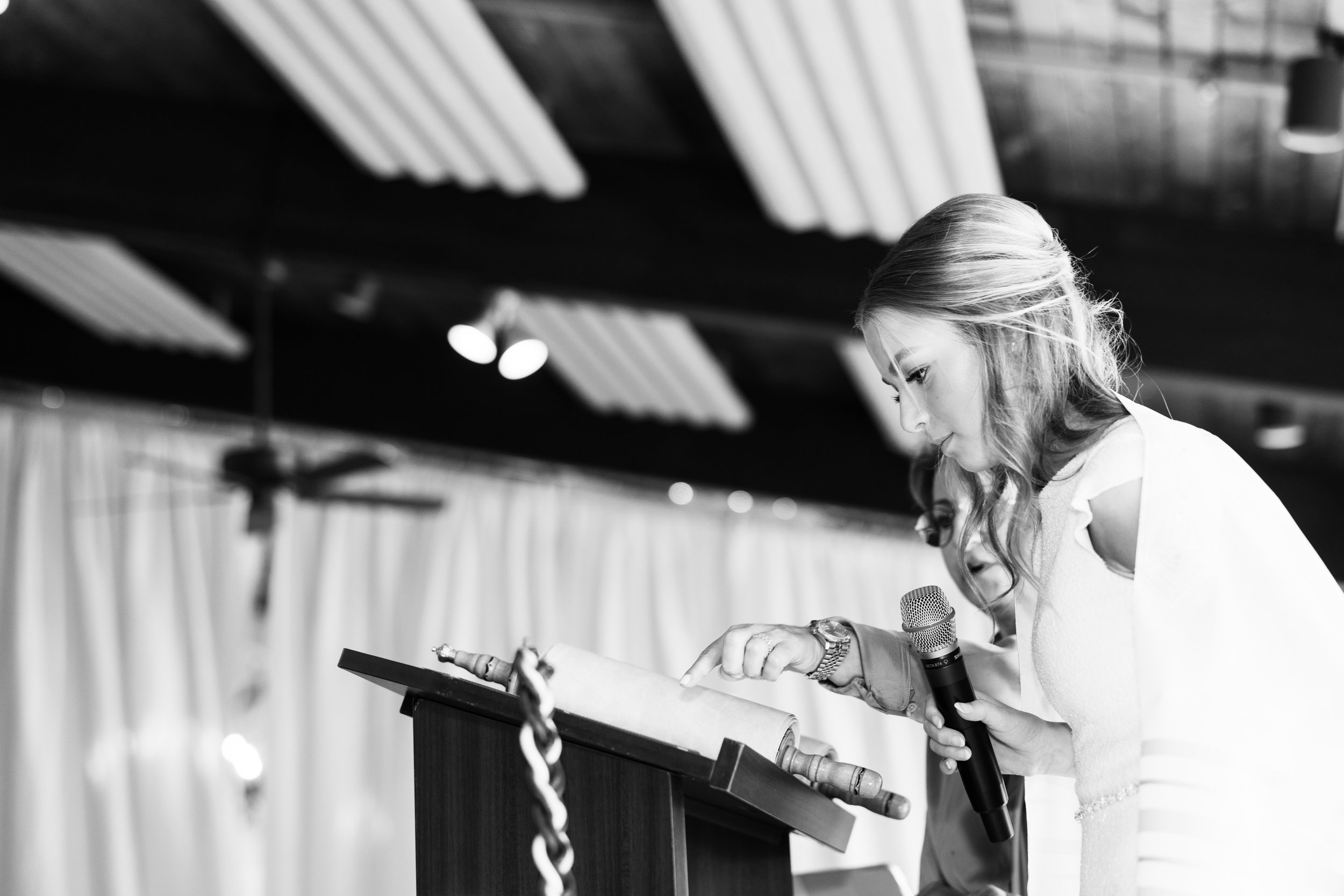 Black and white photo of a woman reading from a book at a podium, holding a microphone, with a woman behind her.