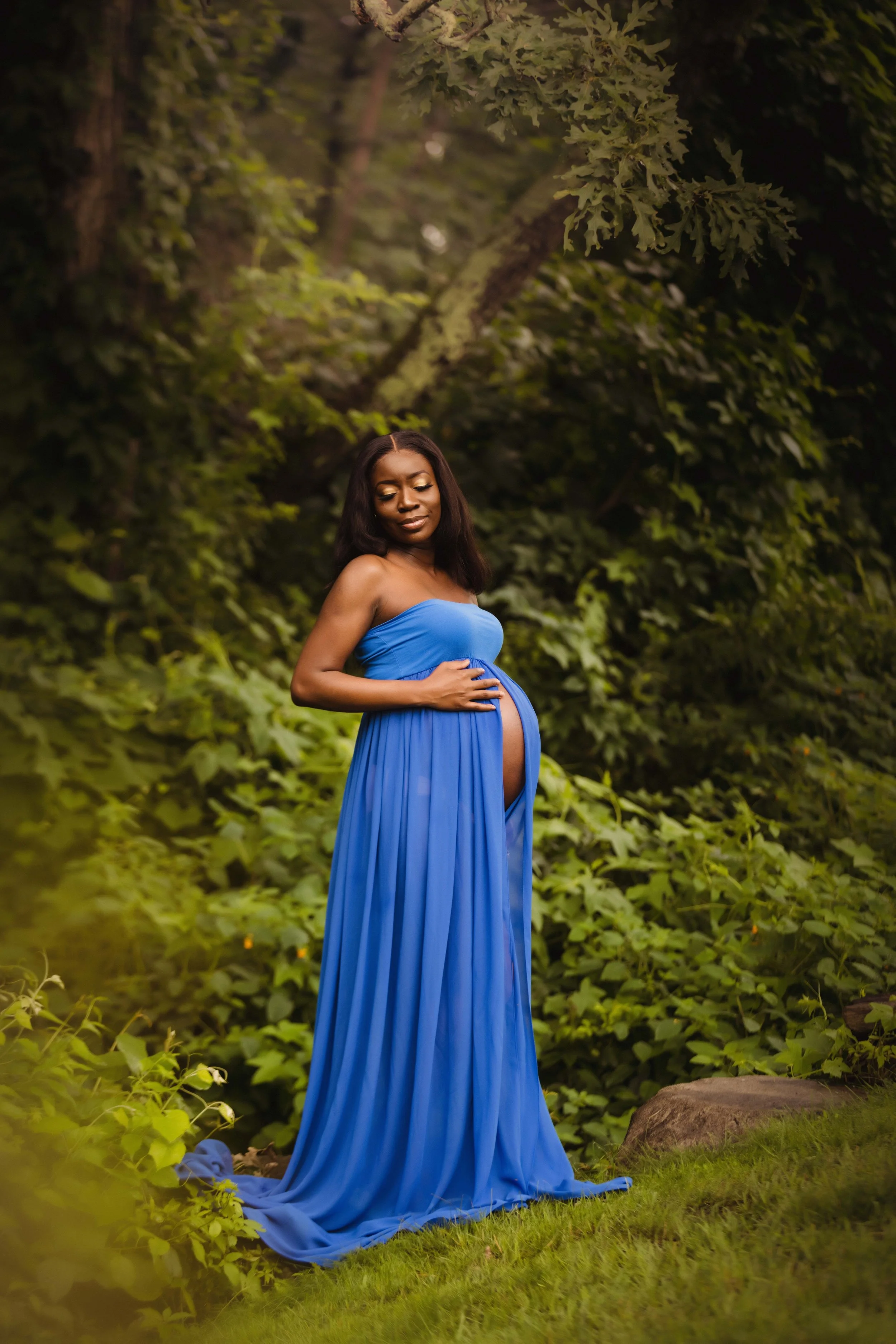 A pregnant woman in a flowing blue dress stands outdoors in a lush green forest, with trees and foliage in the background, gently holding her belly and smiling softly.