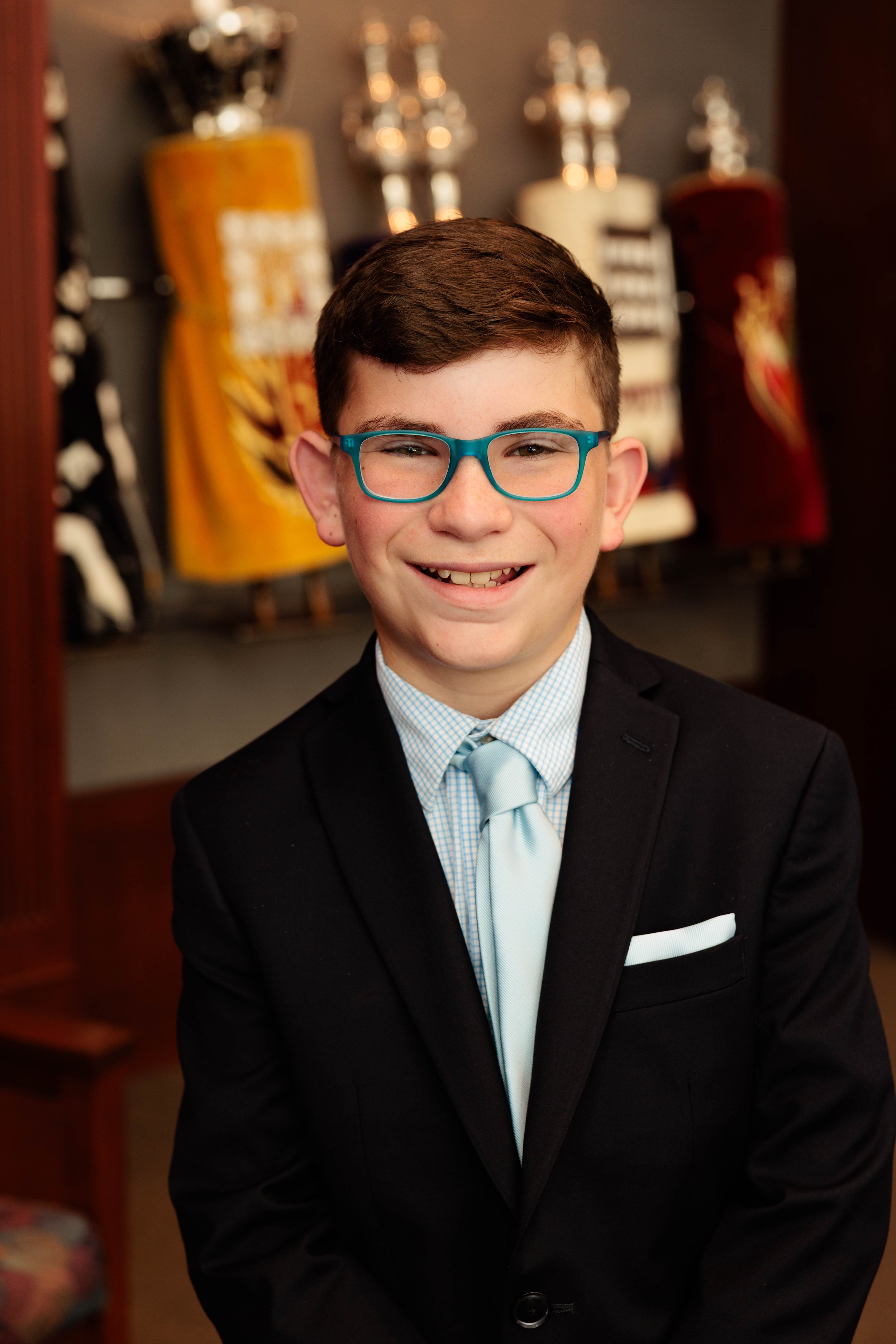 A young boy in a black suit with a light blue tie and glasses, smiling, standing in front of a display of colorful golf-themed jerseys.