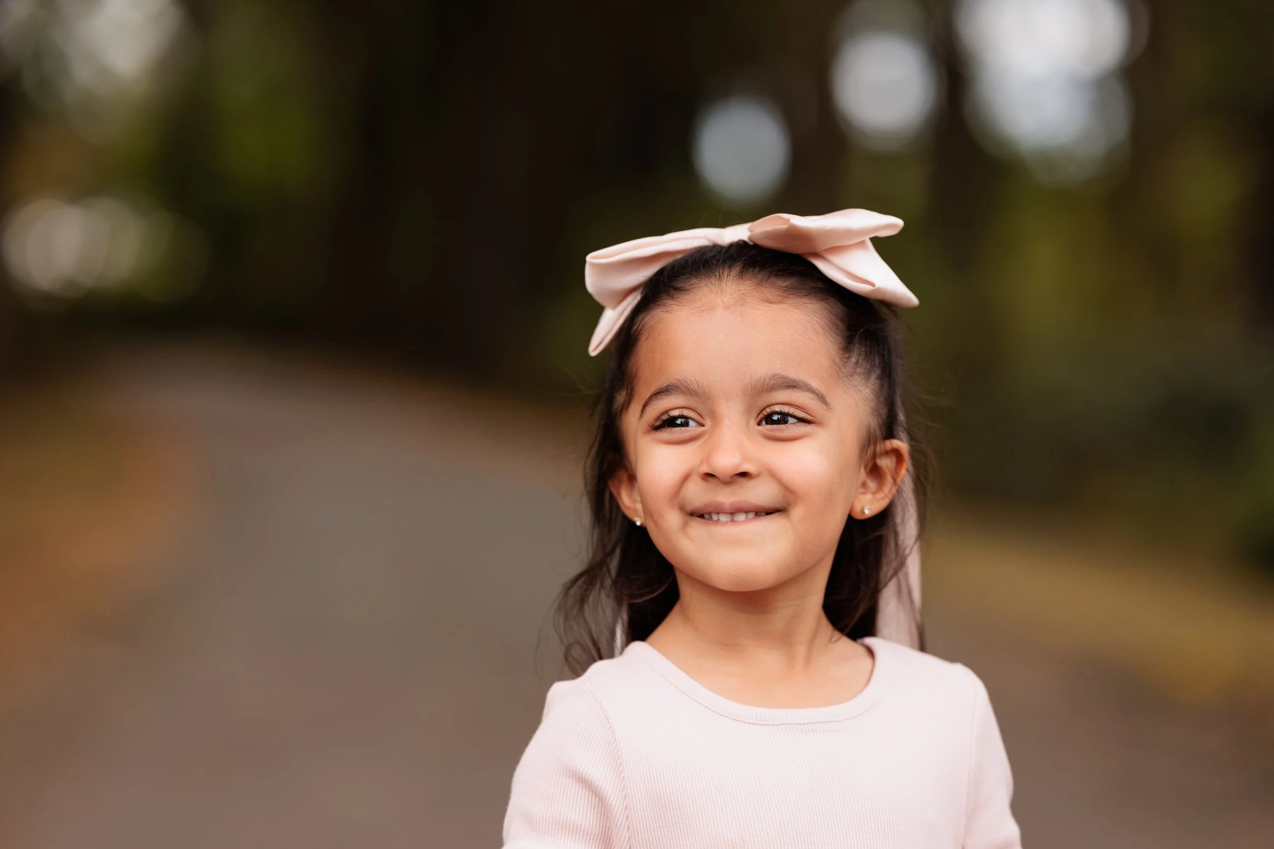 Young girl with a pink bow in her hair smiling outdoors.