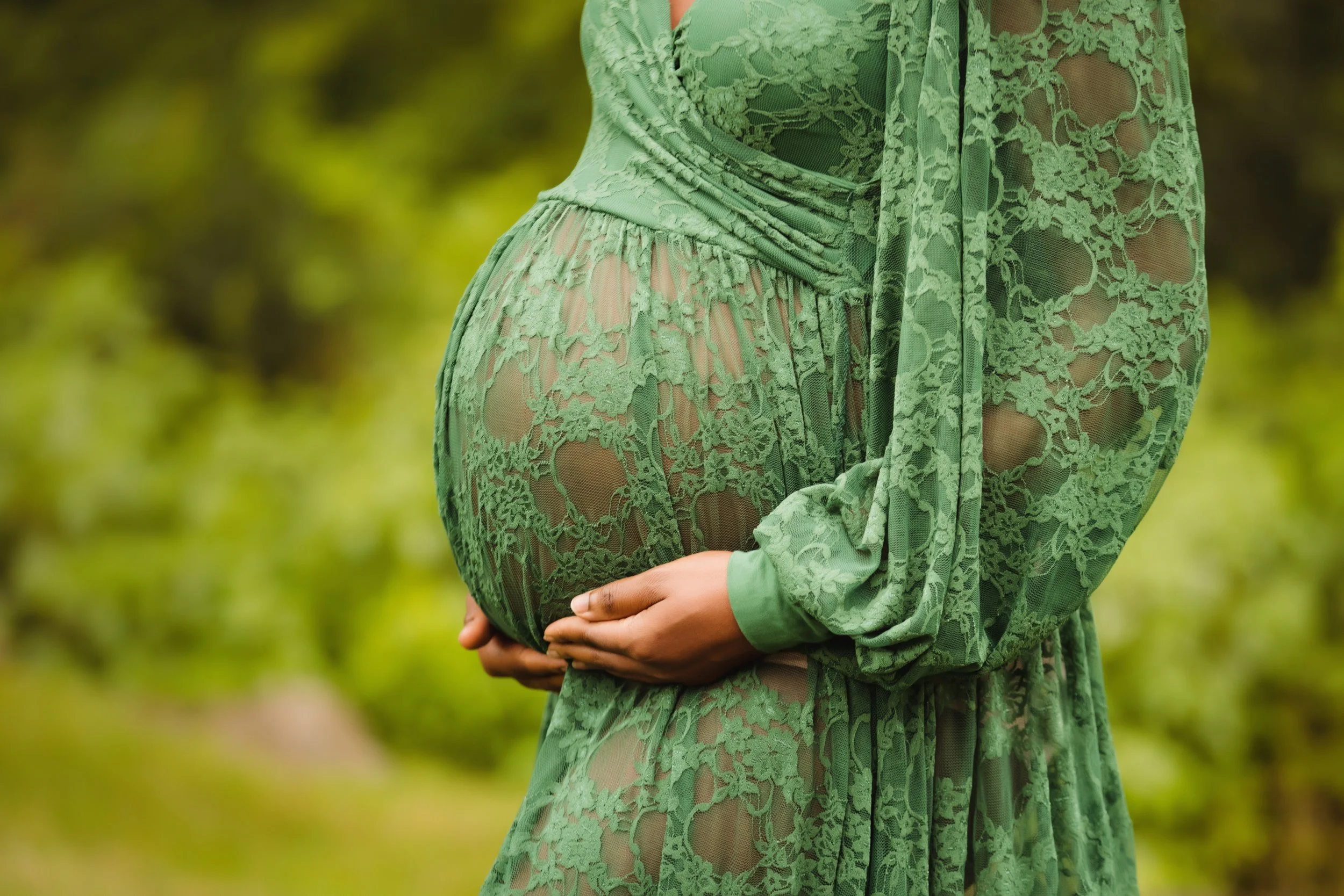 A pregnant woman wearing a sheer green lace dress with floral patterns, standing outdoors in front of a blurred green background, holding her baby bump with both hands.