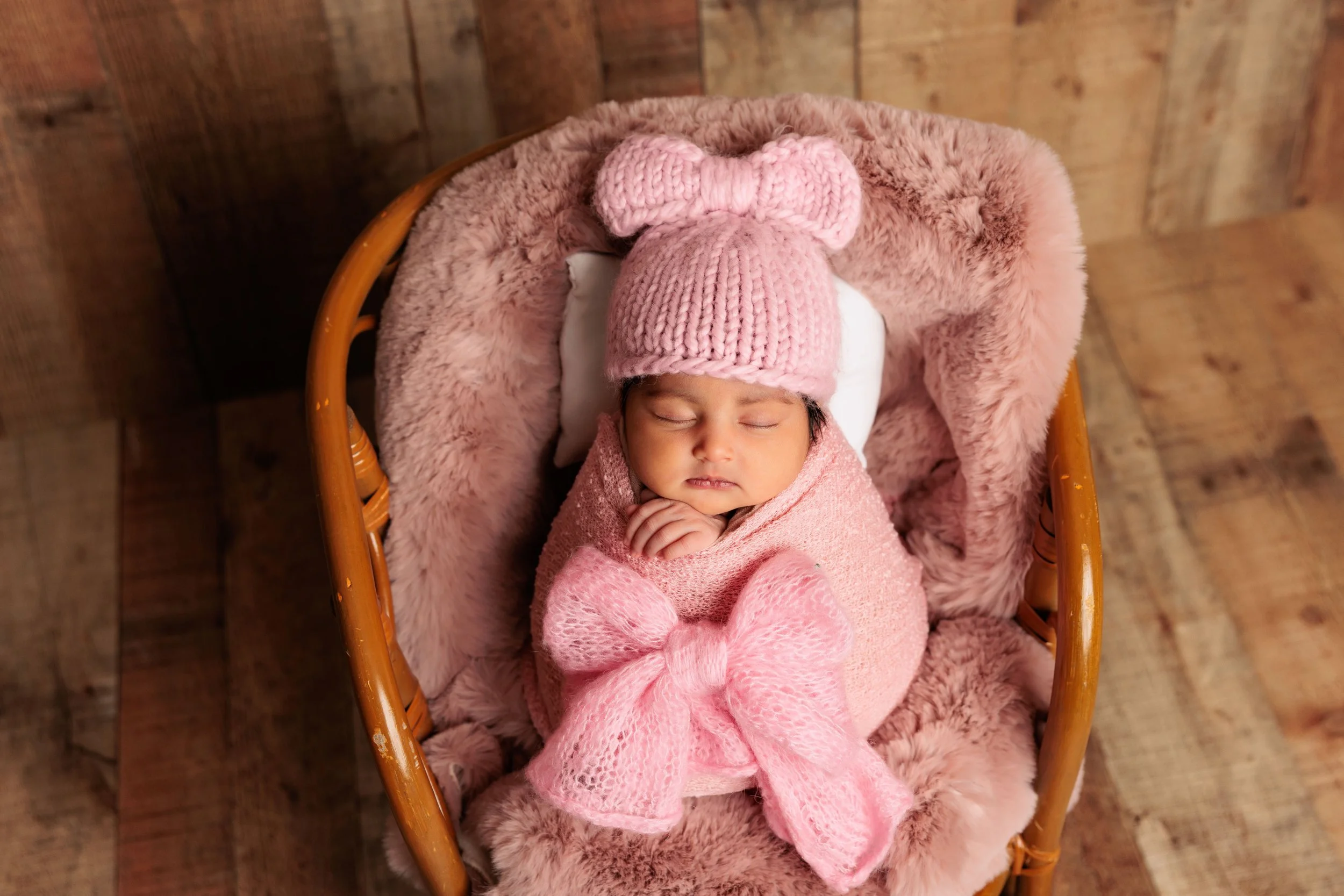 A newborn baby sleeping in a wicker bassinet, wrapped in pink blankets, wearing a pink knitted hat with a bow, and surrounded by soft pink plush material.