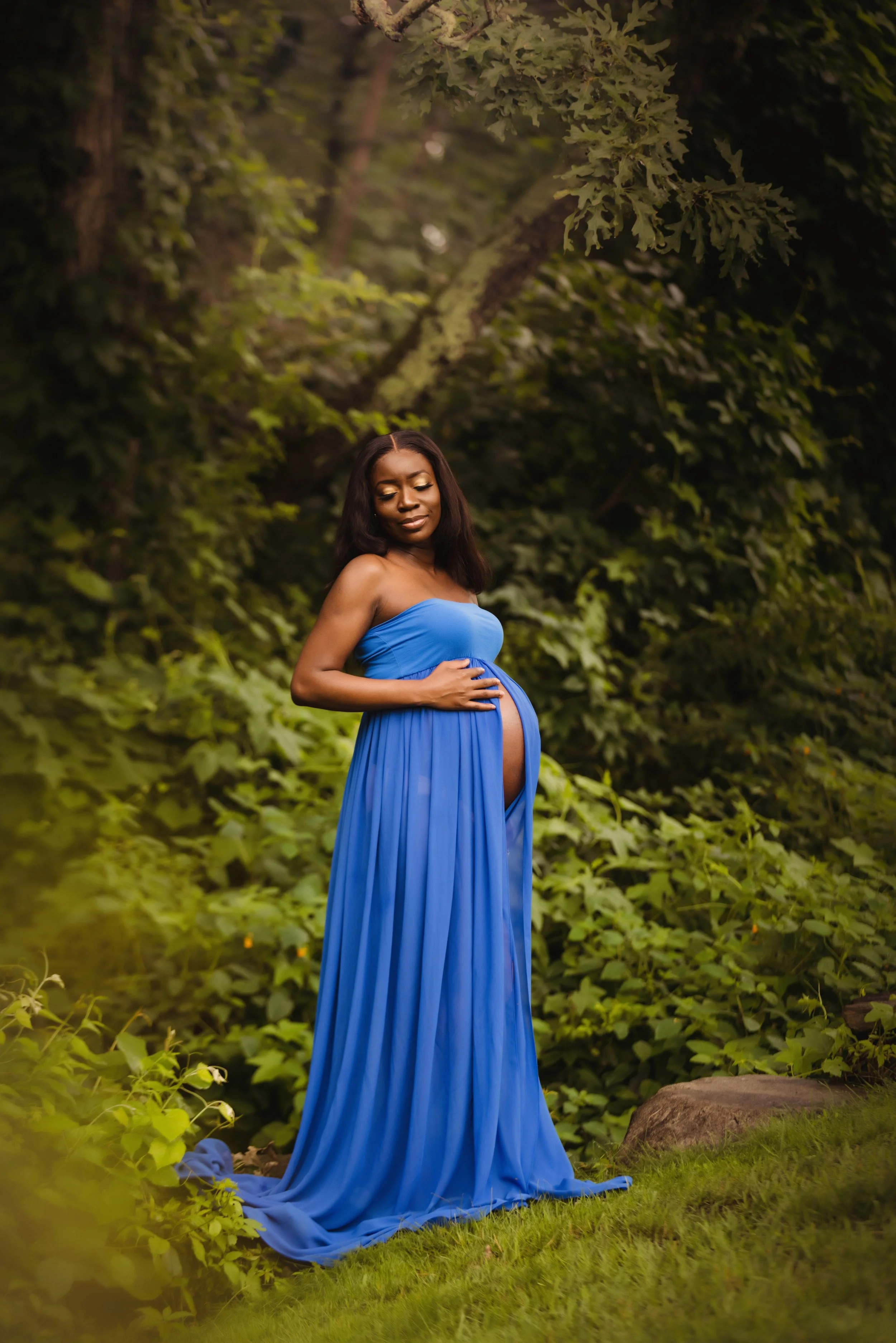 A pregnant woman in a blue strapless gown standing outdoors among green foliage and trees, gently cradling her baby bump and smiling softly.