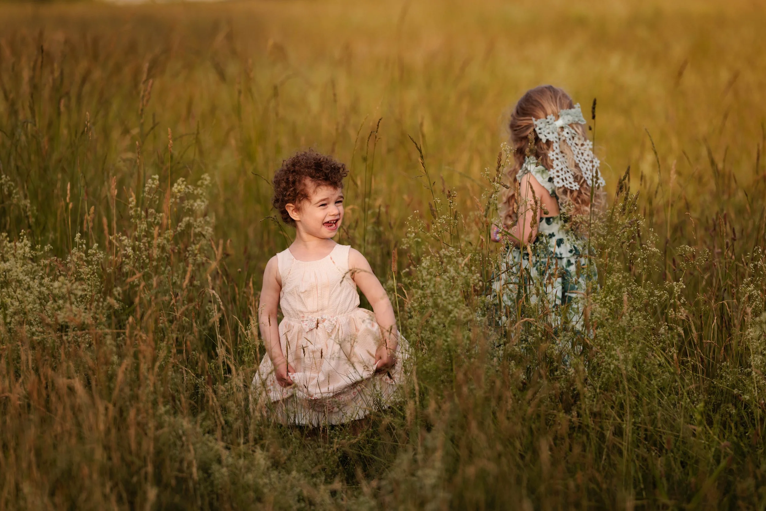 Two young girls standing in a field of tall grass and wildflowers, one with curly hair in a light-colored dress, the other with long wavy hair and a floral headband, enjoying a sunny day.