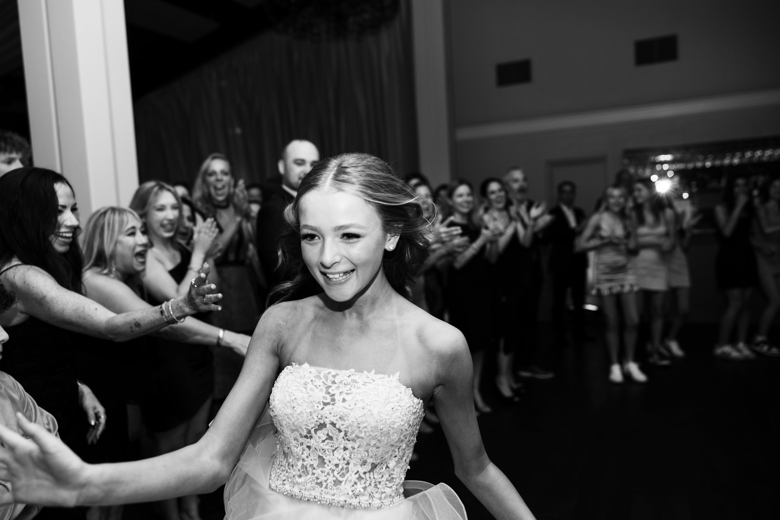 A young woman in a wedding dress smiling while dancing at a celebration with a crowd of people in the background.