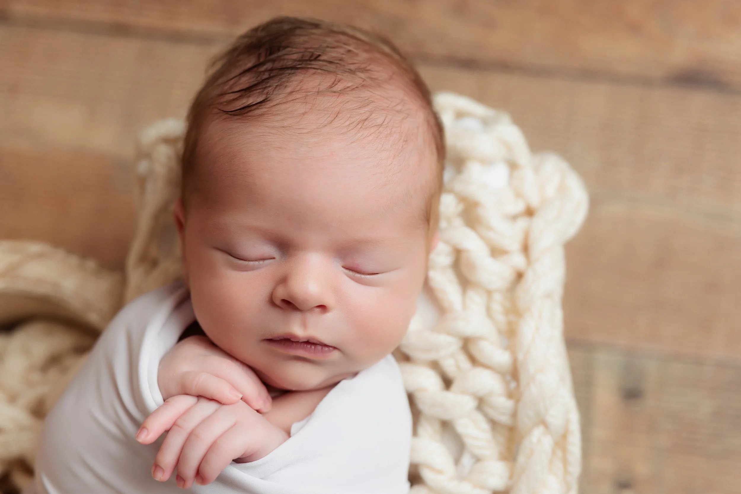 Close-up of a sleeping newborn baby with a hand under chin, lying on a braided blanket on a wooden surface.