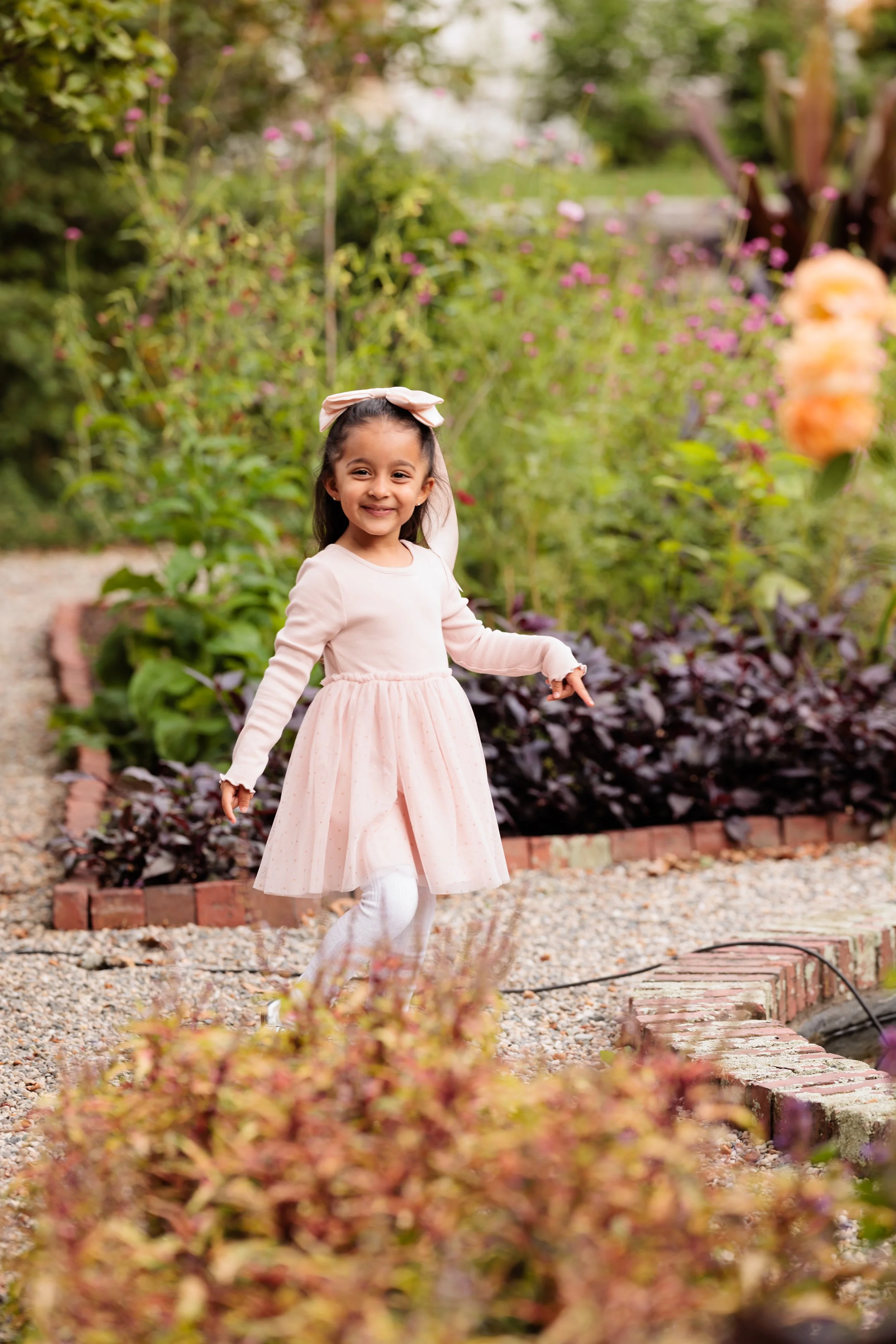 A young girl in a pink dress and white tights smiling and walking in a garden with flowers and greenery.