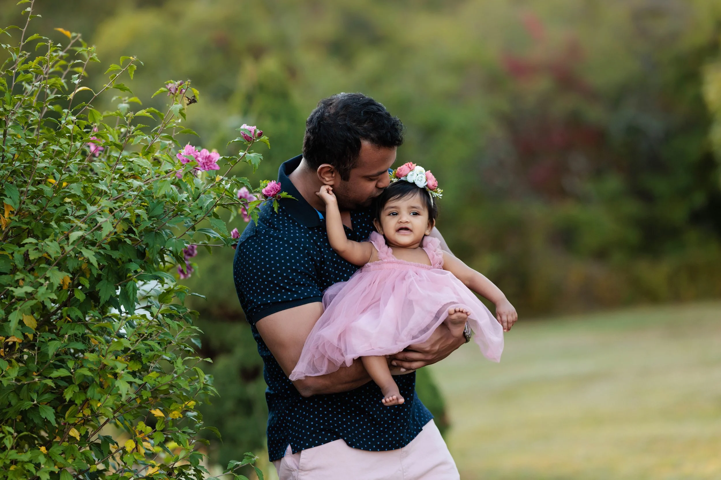 A man holding a young girl in a pink dress and floral headband outdoors near bushes with pink flowers, with a blurred green background.