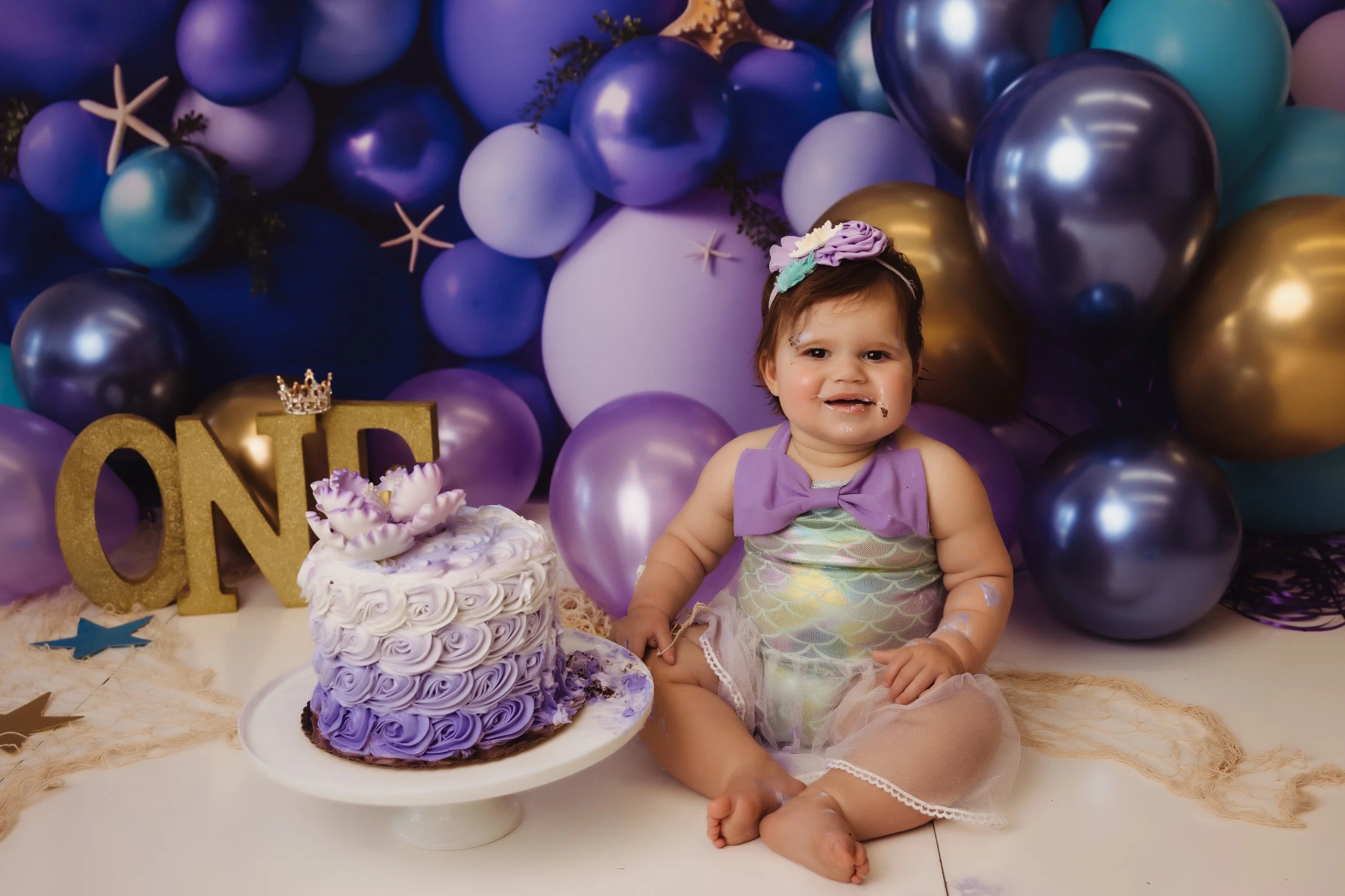 A young girl celebrating her first birthday sits on the floor next to a purple and white birthday cake. She is wearing a shiny, iridescent dress with a purple bow and a floral headband. The background is decorated with purple and gold balloons and st