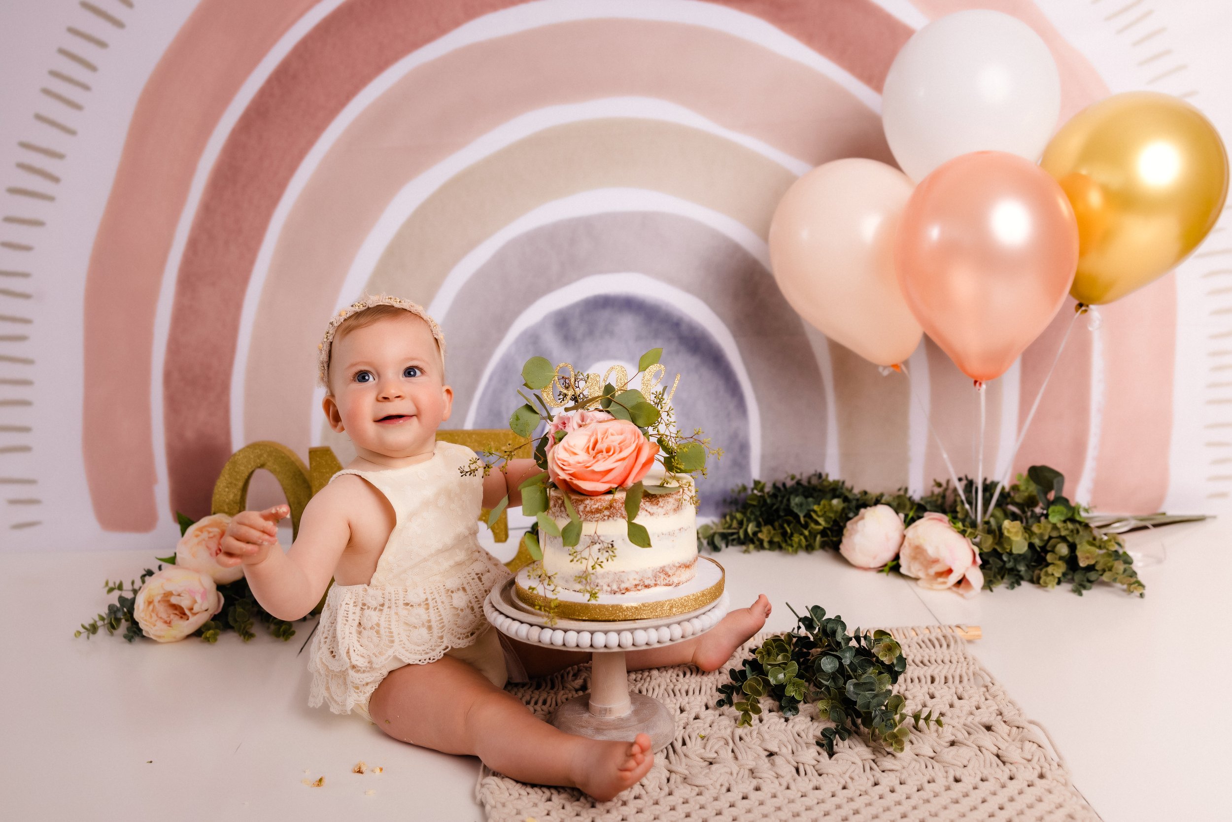 A smiling baby girl in a cream lace dress sitting on a white floor in front of a birthday cake with pink roses and greenery, surrounded by pastel balloons, flowers, and a rainbow backdrop.